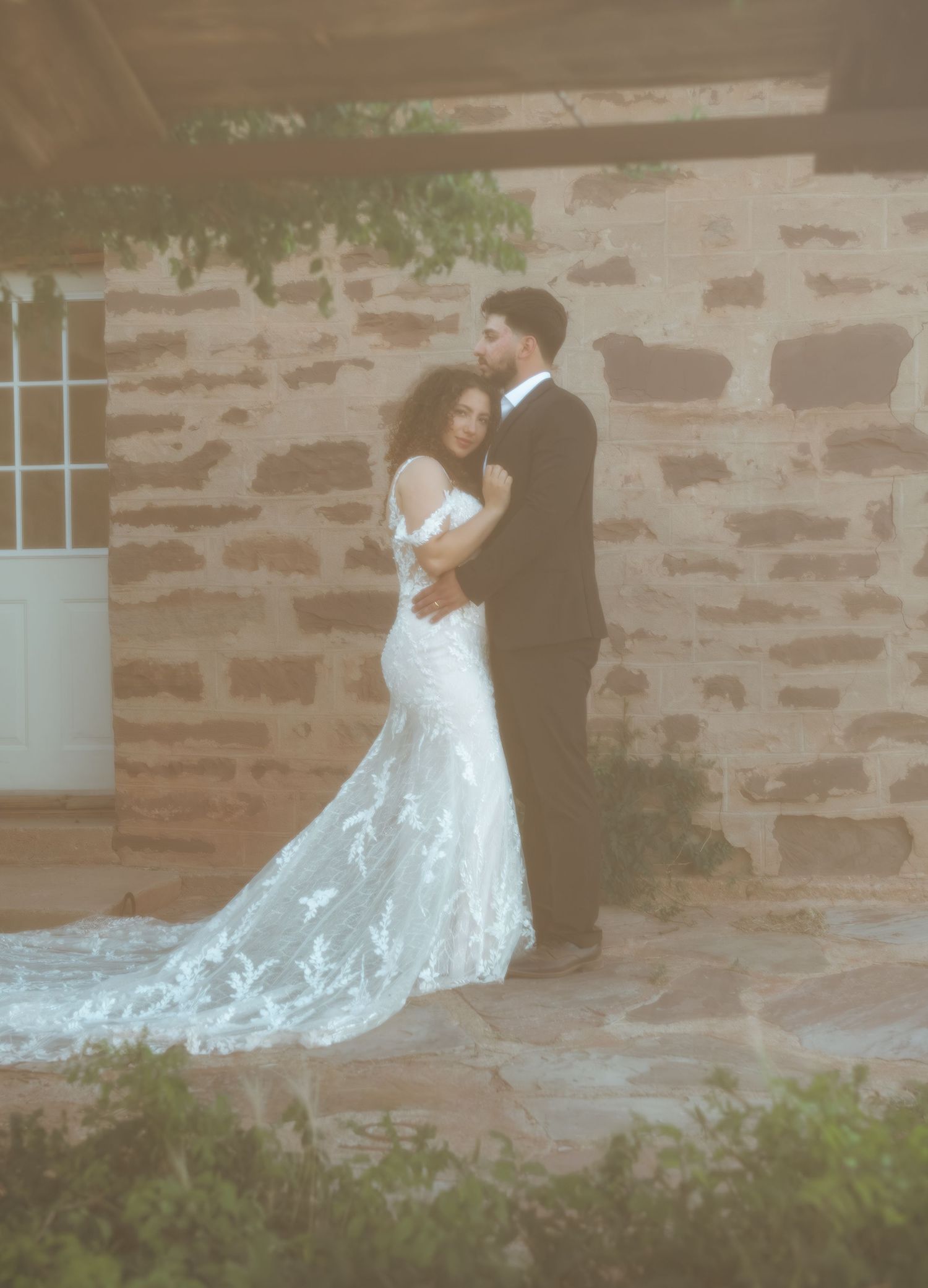Bride in lace mermaid gown and groom in black tux pose by rustic stone wall.