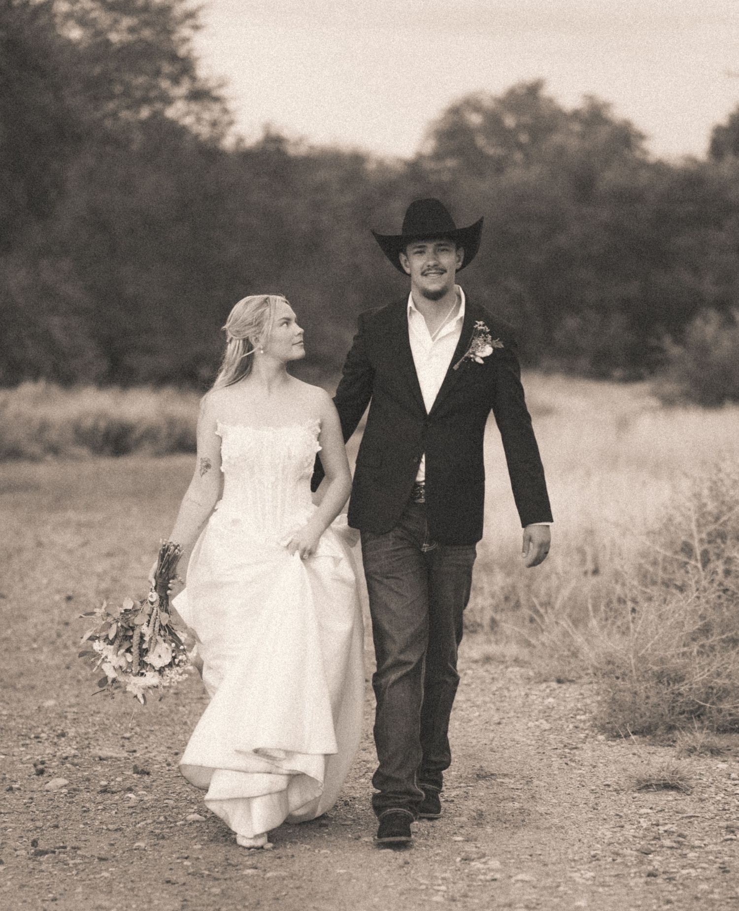 Black and white photo of bride and groom walking hand in hand on dirt path.
