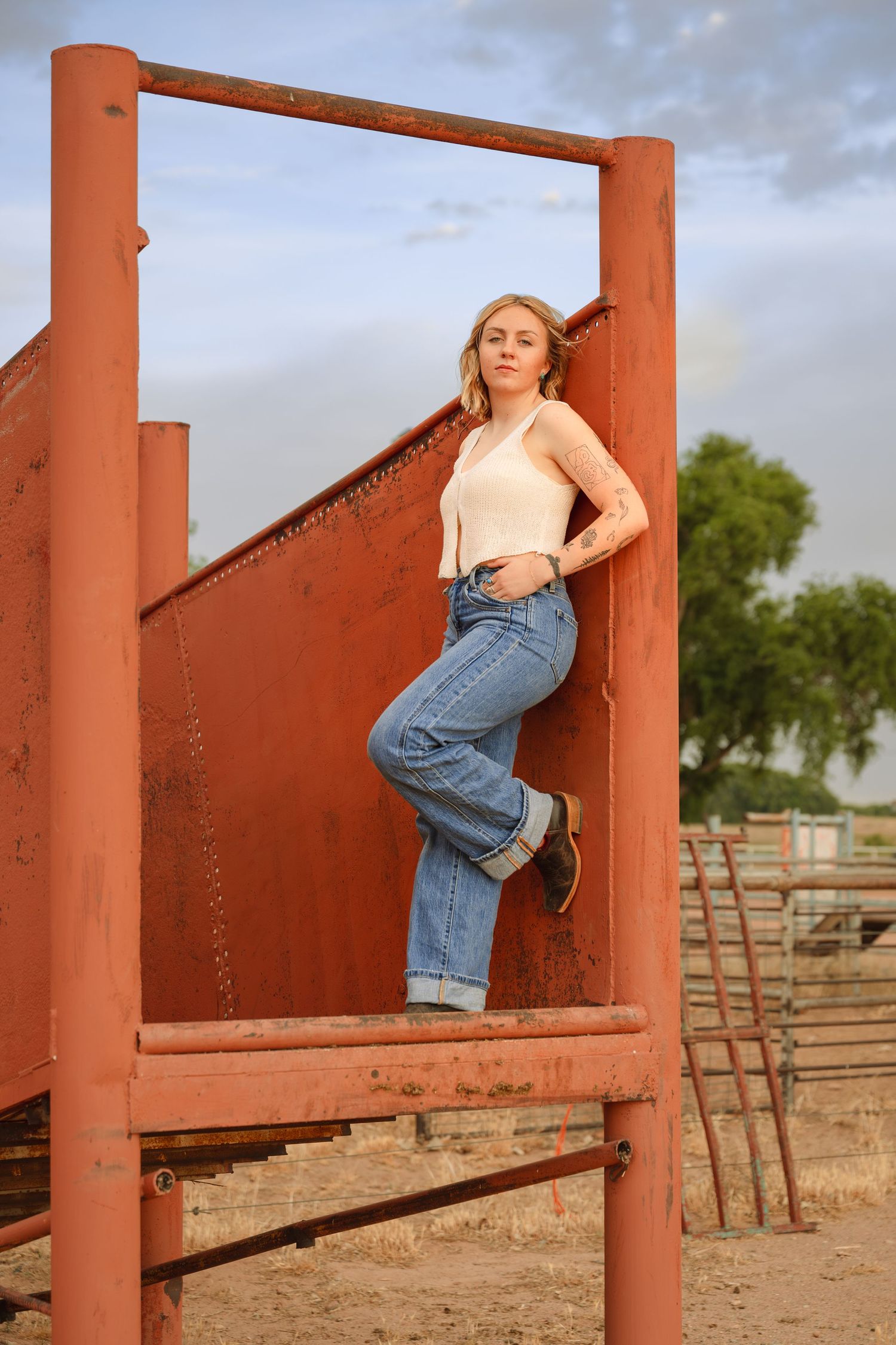 Young blonde woman in jeans and white top posing on rustic red metal farm equipment outdoors.