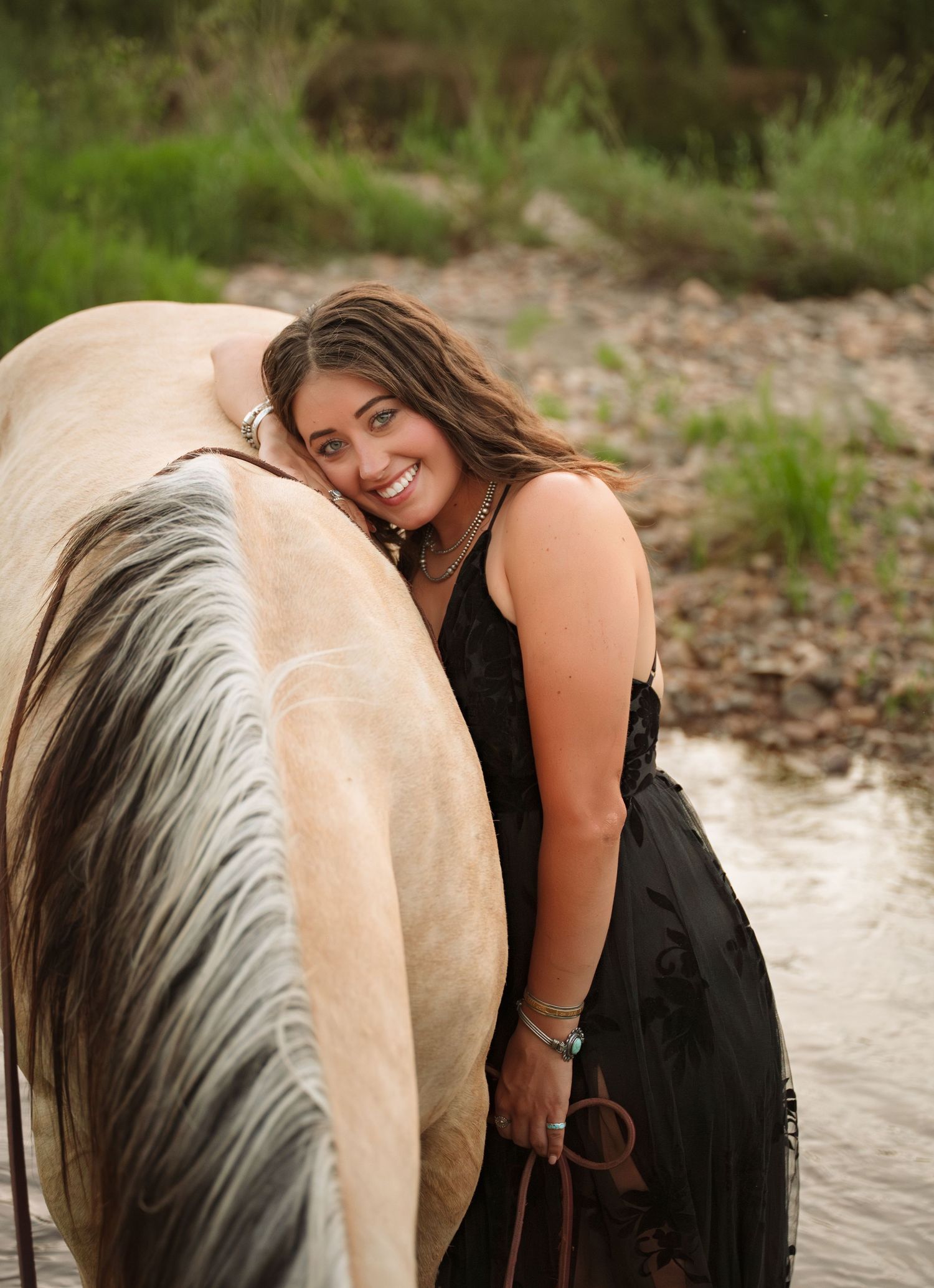 A smiling young woman in a black dress leans affectionately against a pale horse near a creek with green trees.