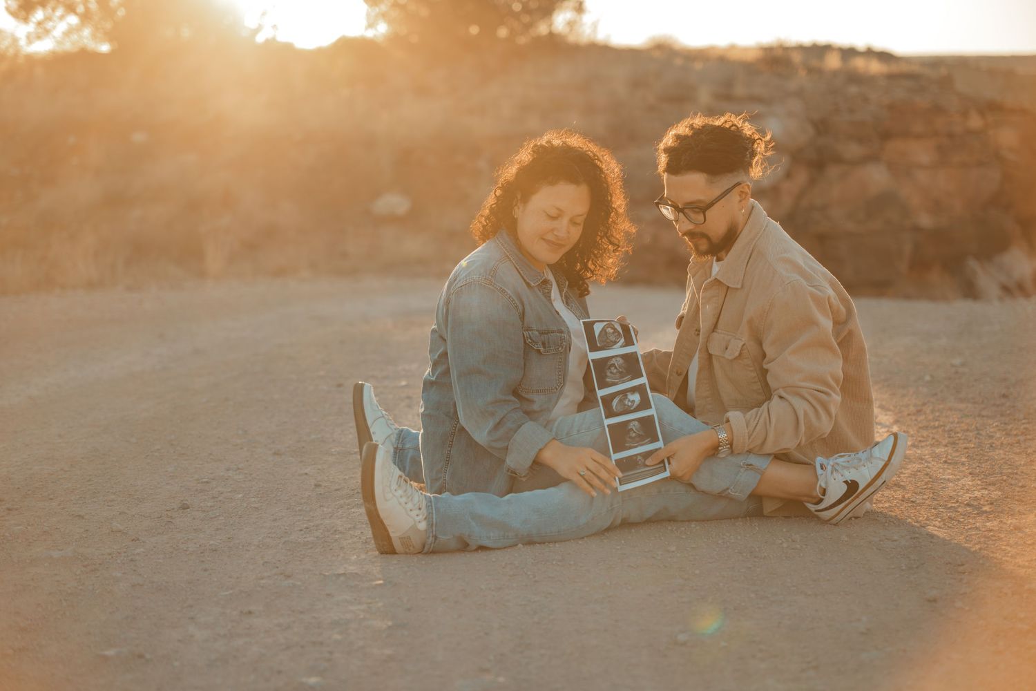 Couple sitting in desert at sunset holding ultrasound photos, announcing pregnancy in golden light.