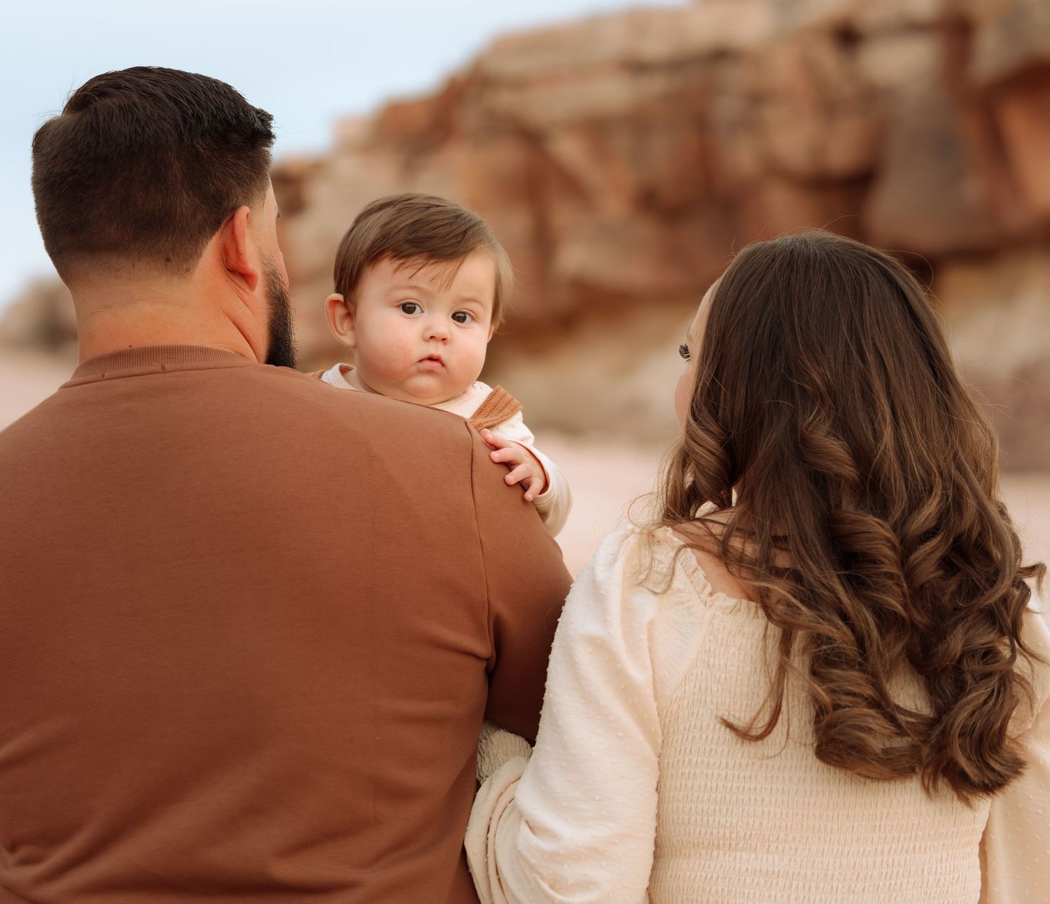 Parents holding baby facing red rock formations, baby looking back at camera.
