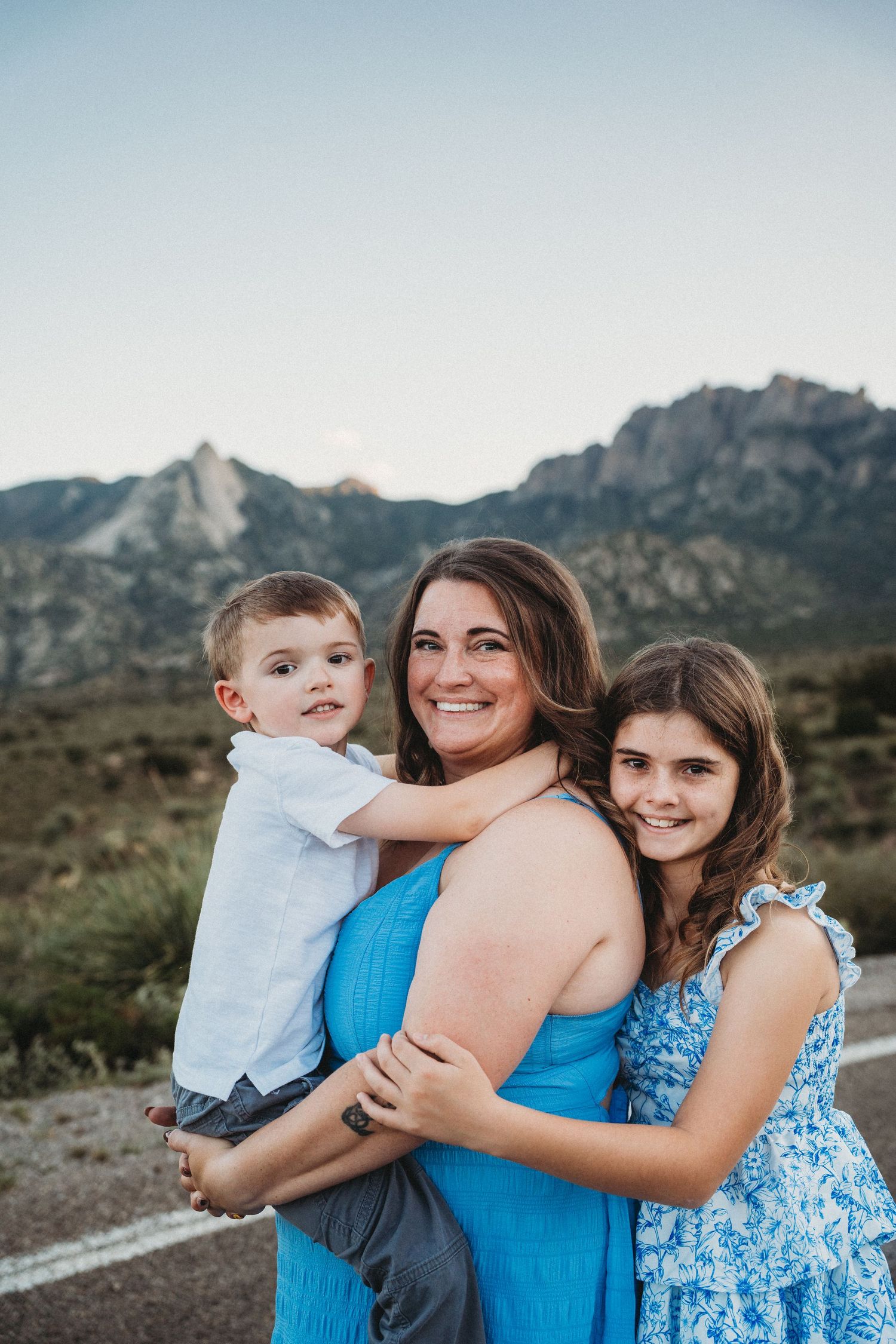 Smiling mother in blue dress holding young son while daughter leans in against mountain backdrop.