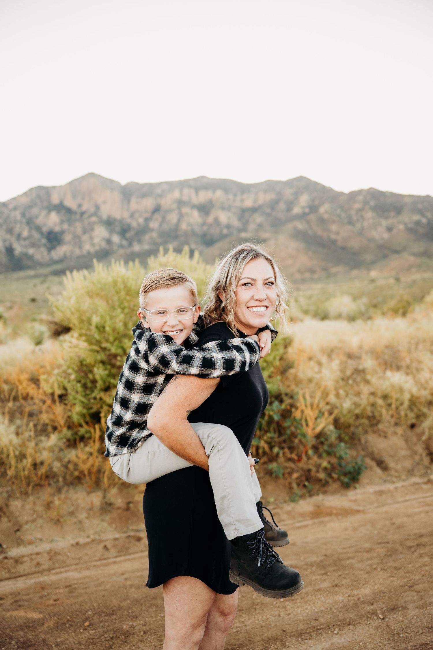 A smiling woman gives a young boy a piggyback ride on a scenic desert mountain trail.
