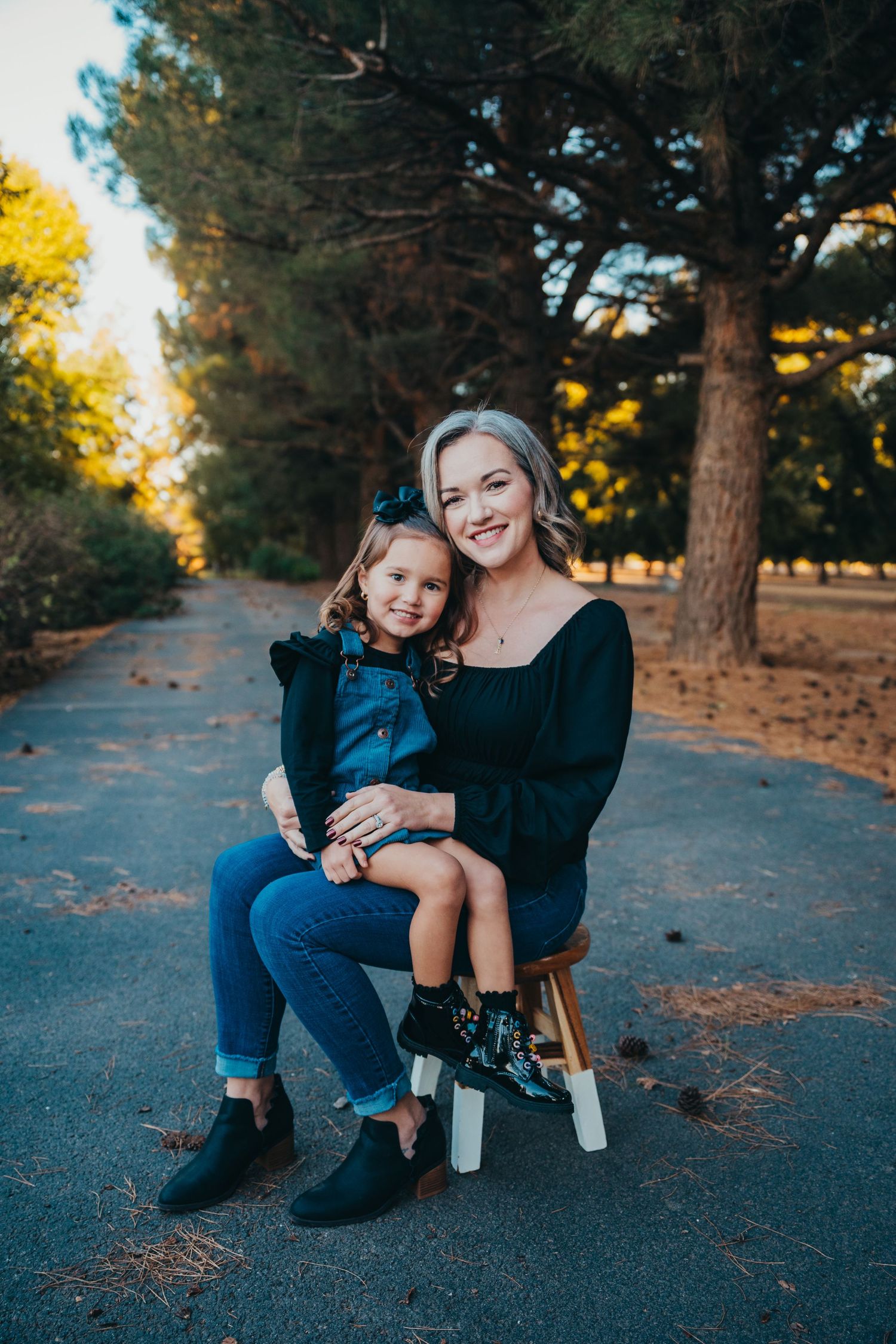 Mother in black top sitting on a stool with young daughter in denim overalls on her lap outdoors.