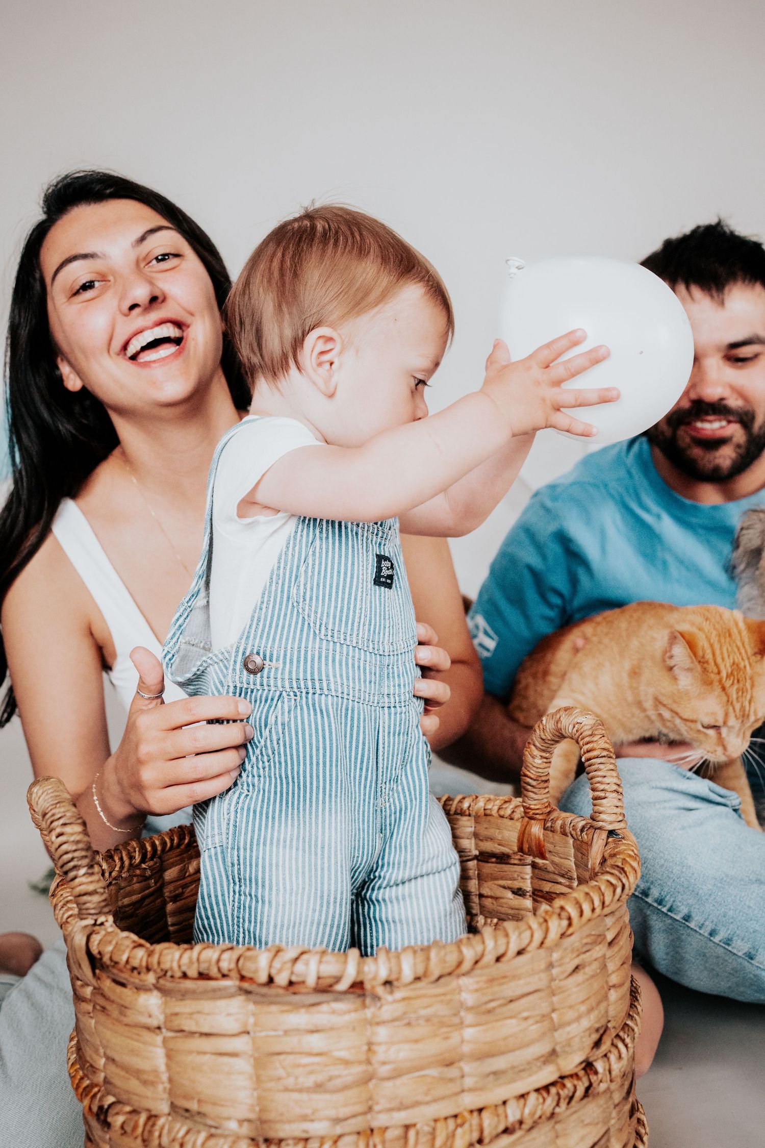 Laughing mother holding baby in wicker basket while father pets orange cat nearby.