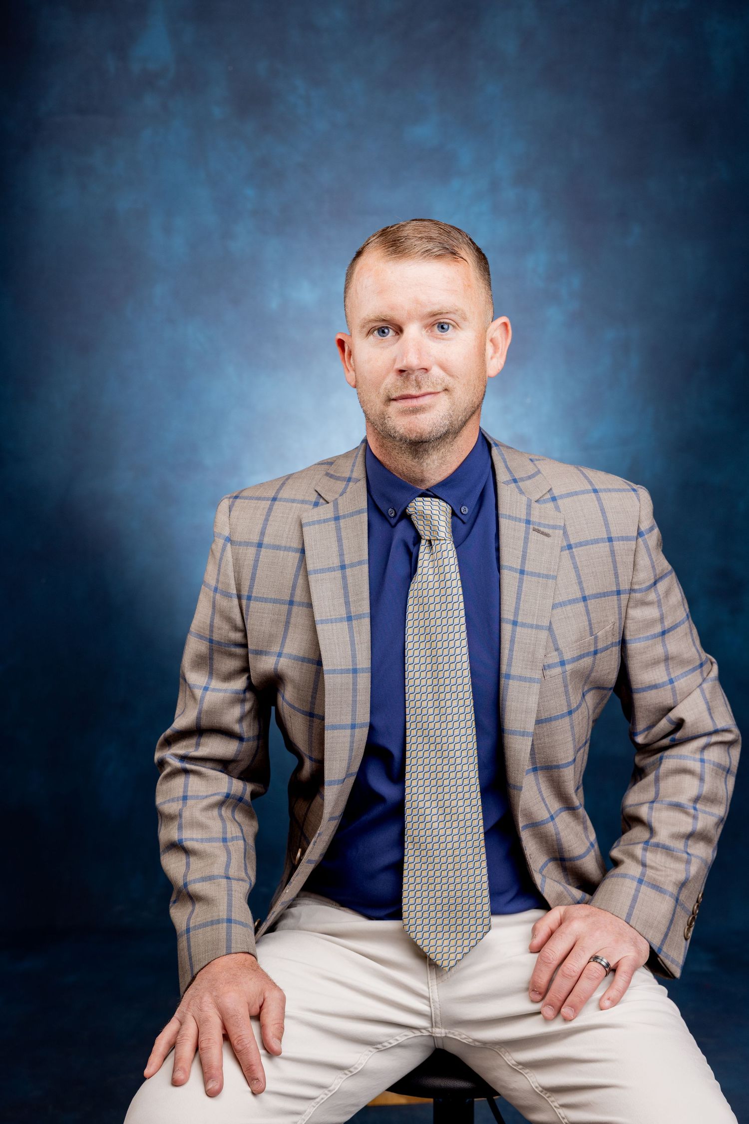 Full-length business portrait of a man in a plaid blazer and blue shirt seated against a dark blue backdrop.