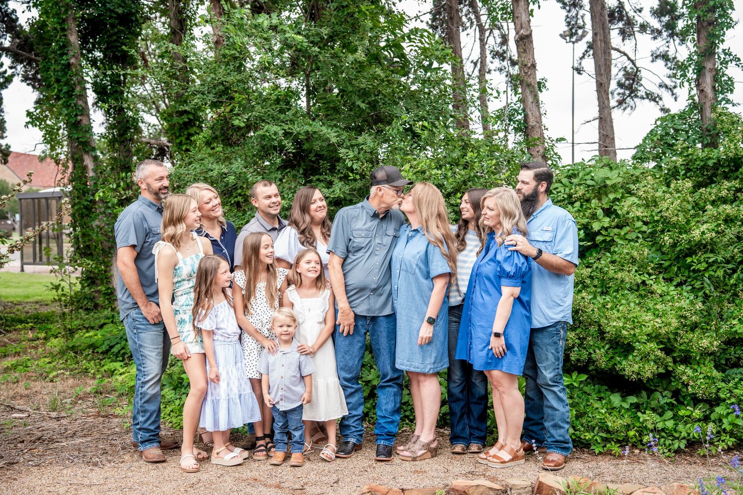 Large multigenerational family group portrait outdoors surrounded by lush green trees and foliage.