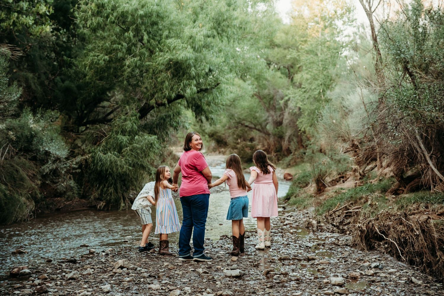 Mother walks hand-in-hand with four children along a tree-lined creek bank.