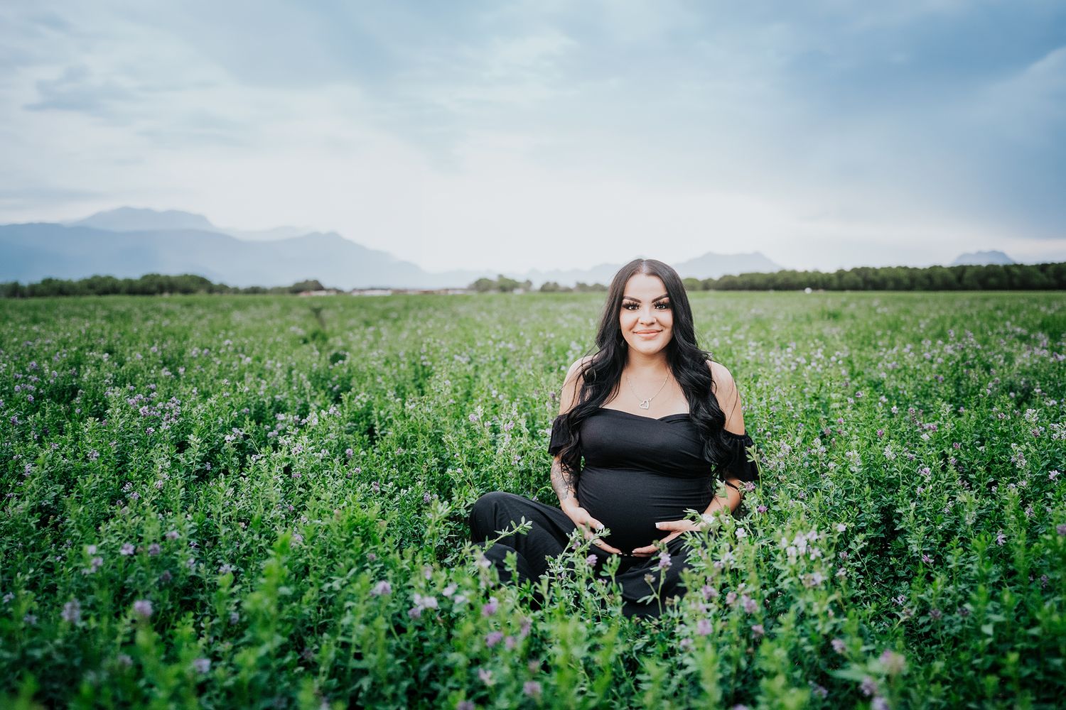 Pregnant woman in black outfit sitting in lush purple wildflower field with mountains.