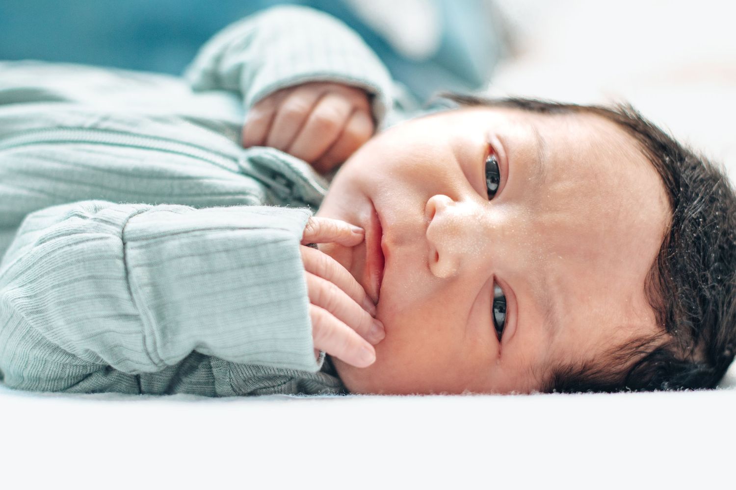 Close-up of an awake newborn baby lying down, hand near face, wearing a light green outfit.