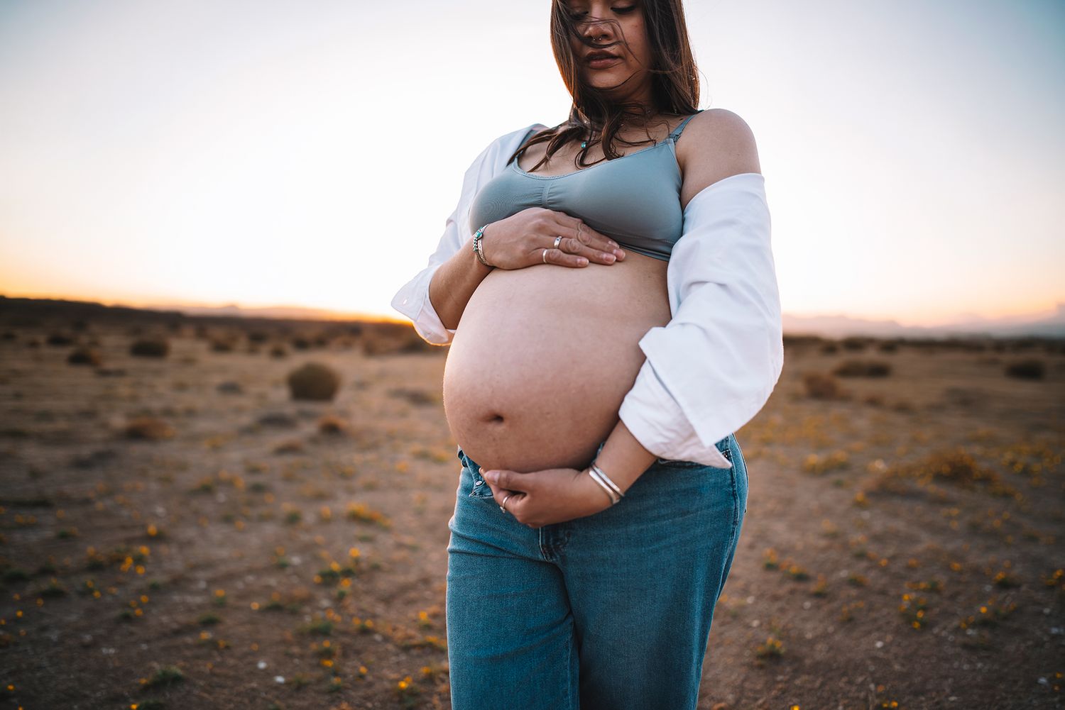 Close-up of pregnant woman holding bare belly in desert wearing jeans at sunset.