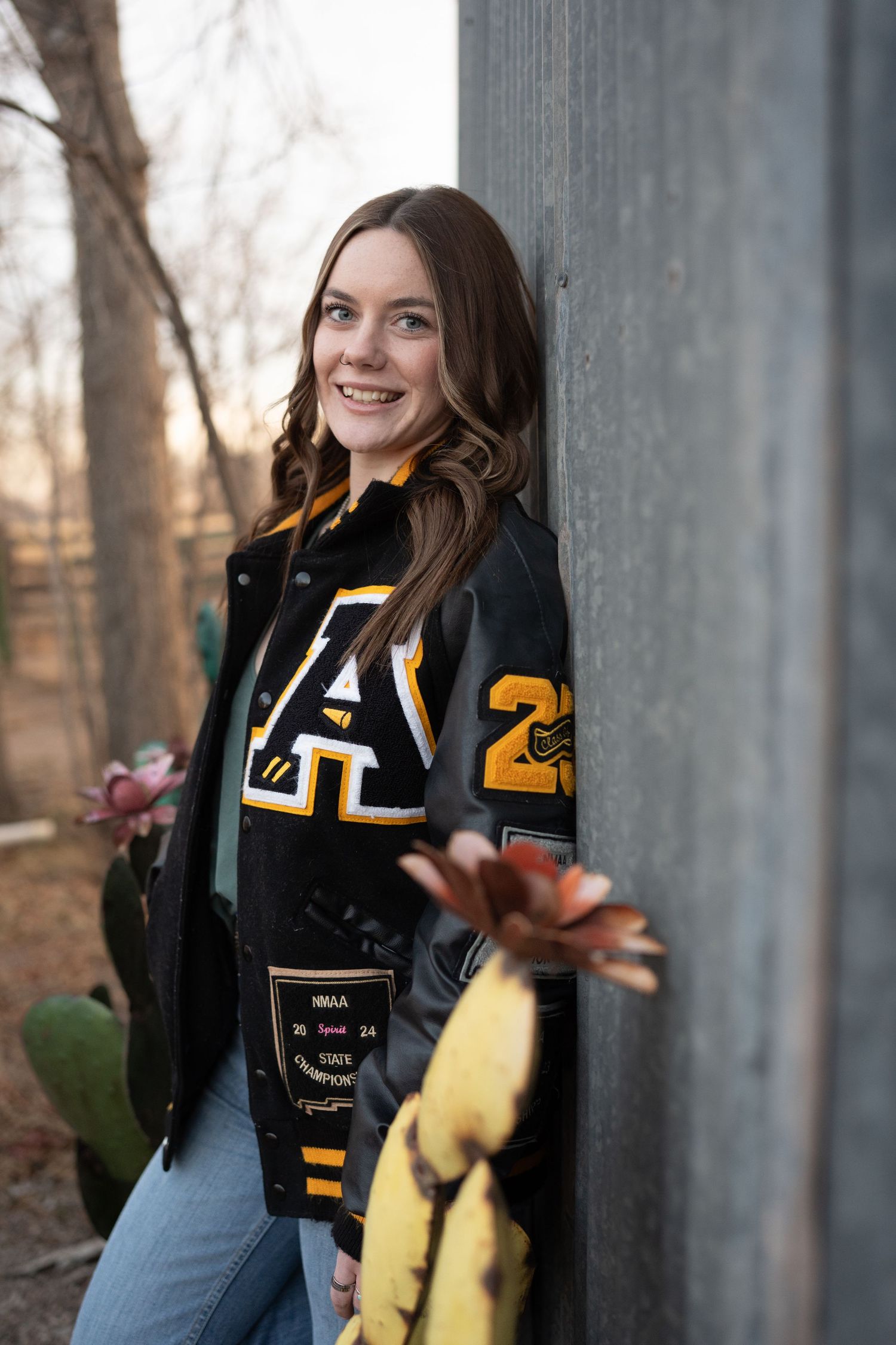 Smiling senior girl in black and gold letterman jacket leans against gray wall outdoors.