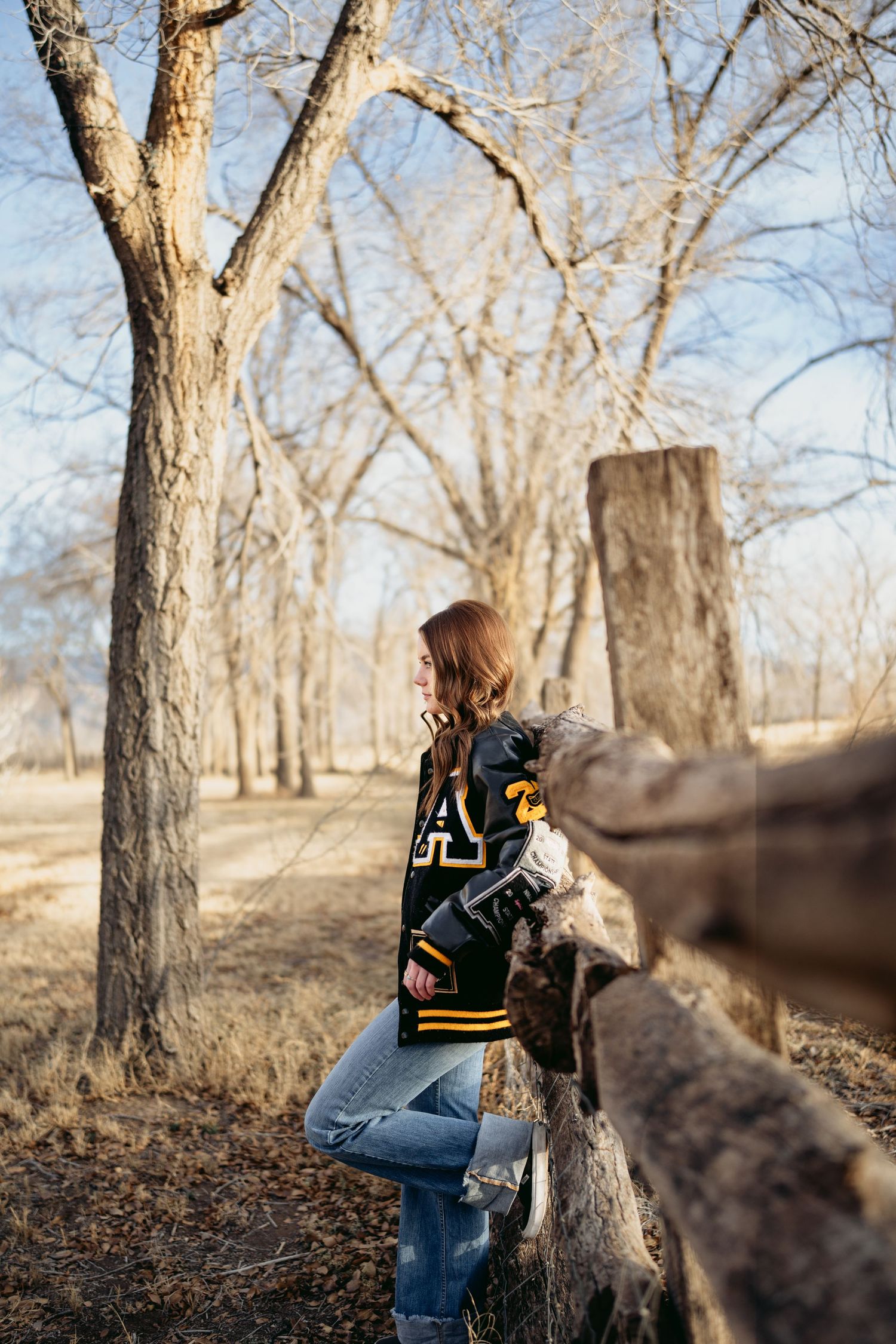 Young woman in black and gold letterman jacket leaning on rustic wooden fence among bare winter trees.