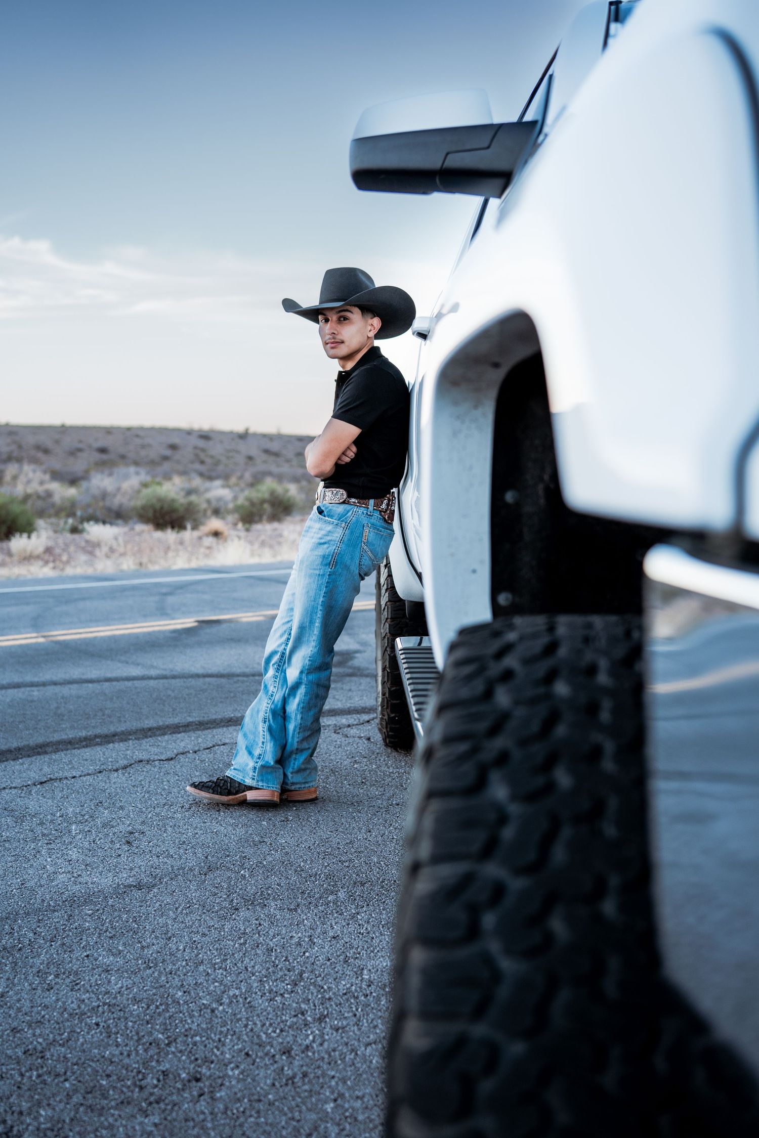 Young man in black cowboy hat and jeans leaning against white GMC truck on desert highway.