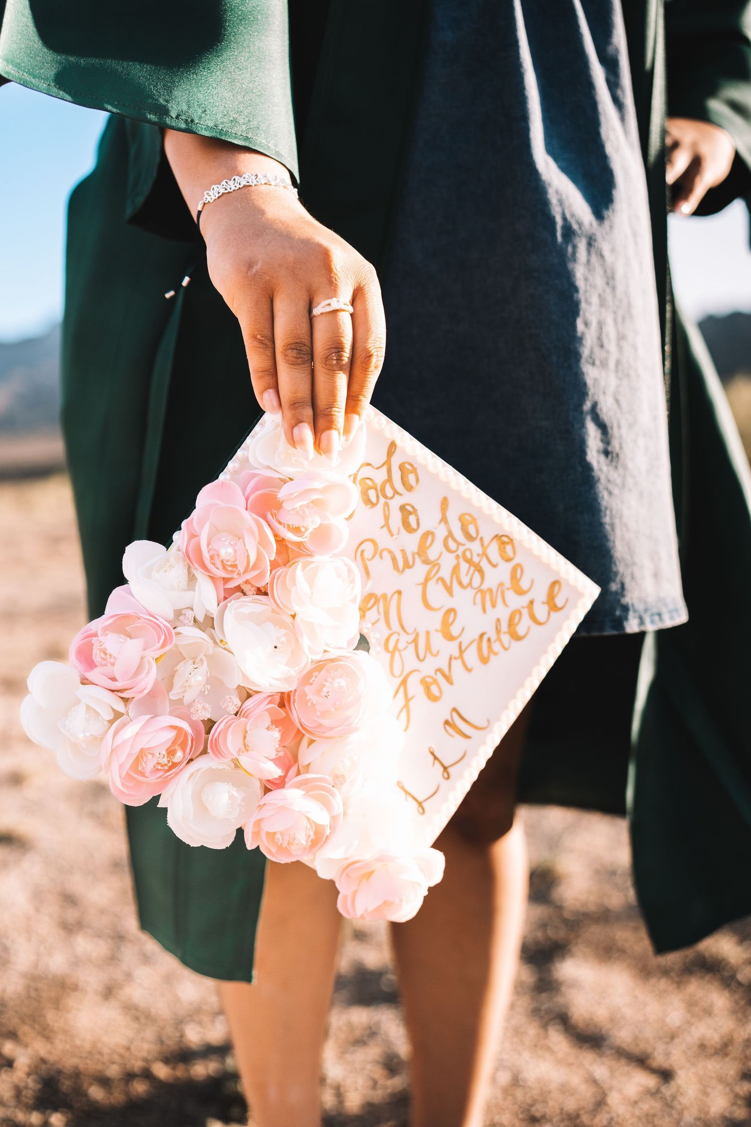 Graduate holding a decorated cap with pink flowers and inspirational Spanish quote outdoors.
