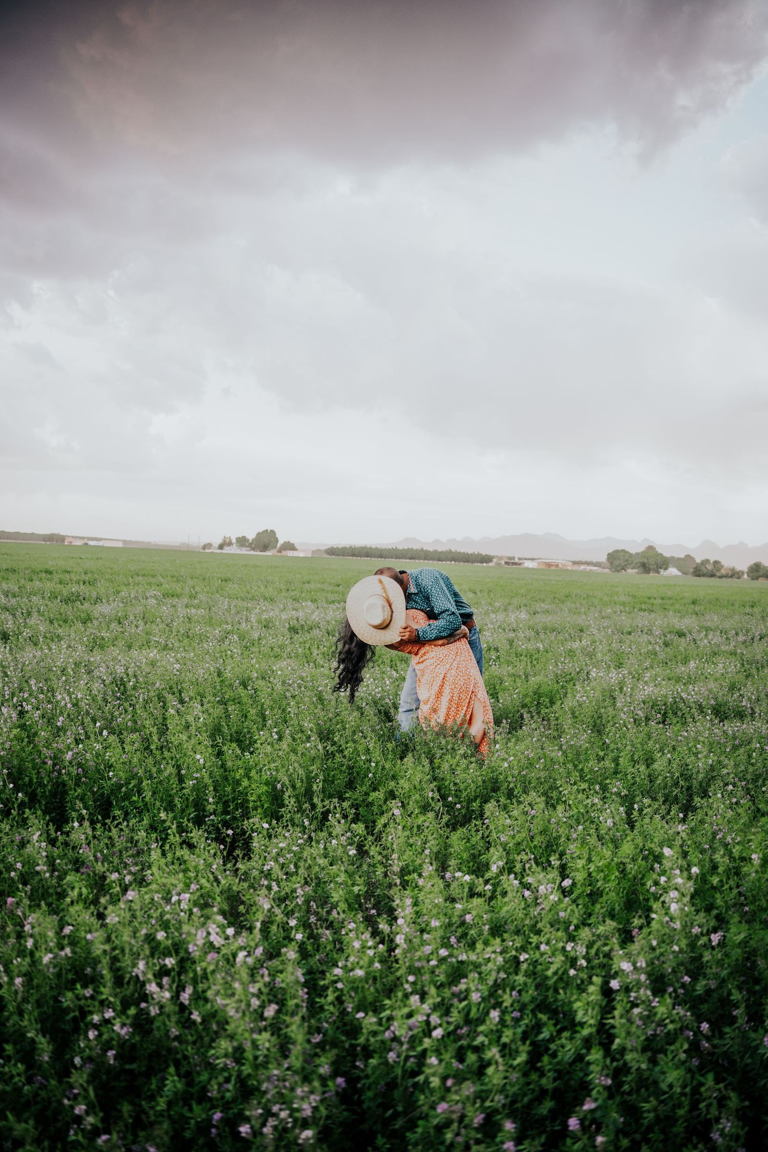 Couple leaning together in a green field, man in cowboy hat and teal shirt, woman in orange floral dress.
