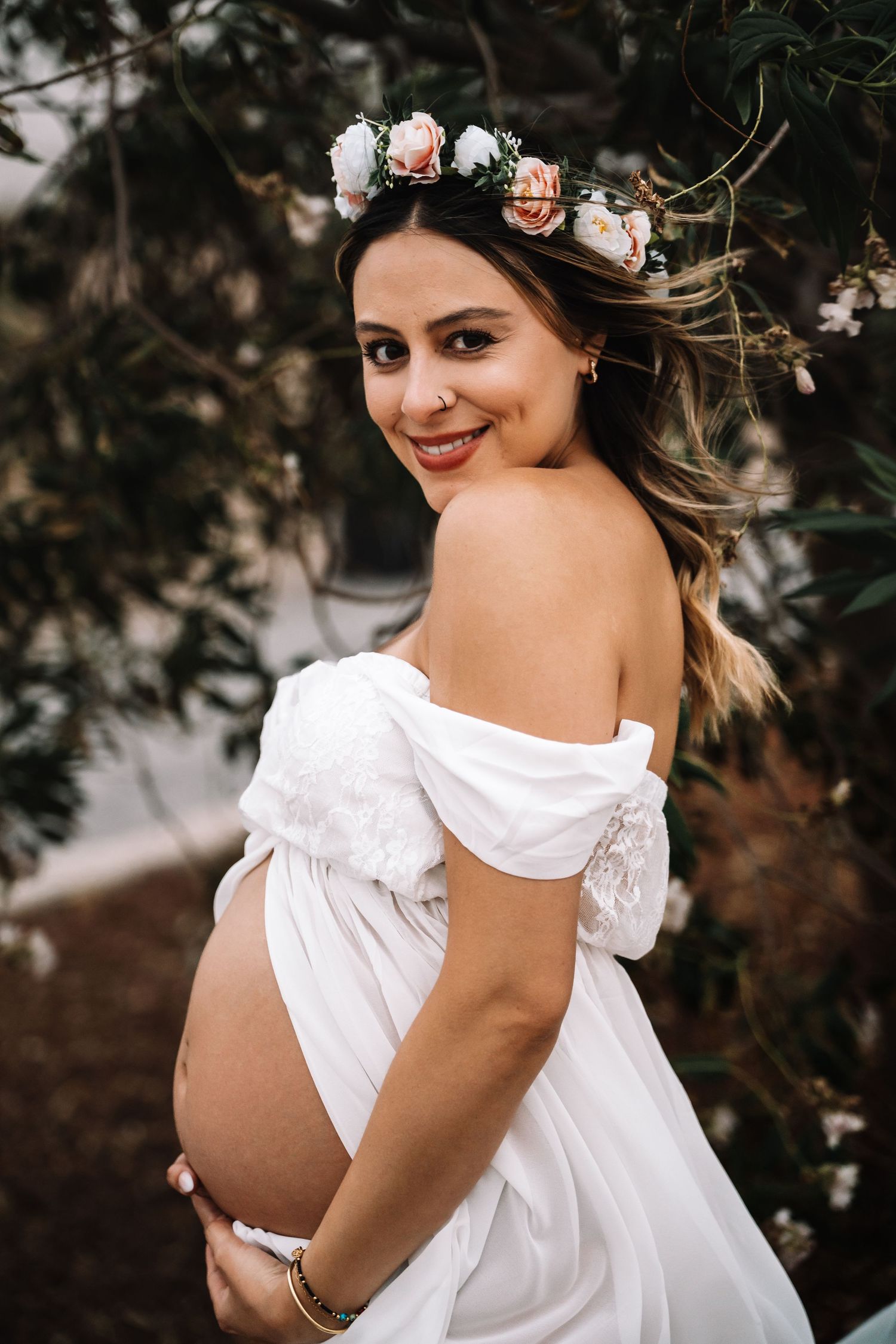 Smiling pregnant woman wearing floral crown and white off-shoulder maternity dress poses among garden blooms.