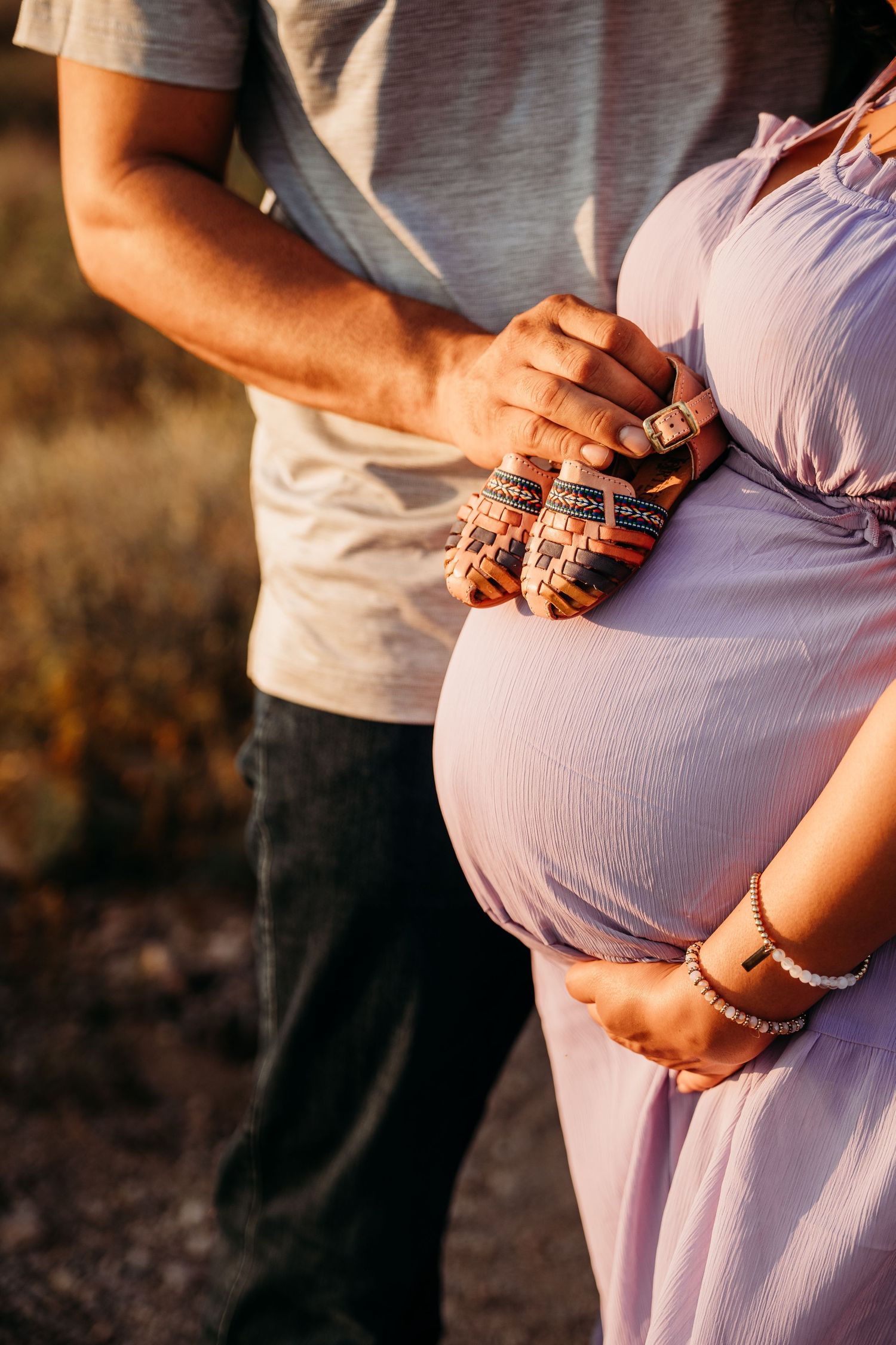 Couple cradling baby booties against pregnant belly in purple dress, bathed in warm golden hour sunlight.