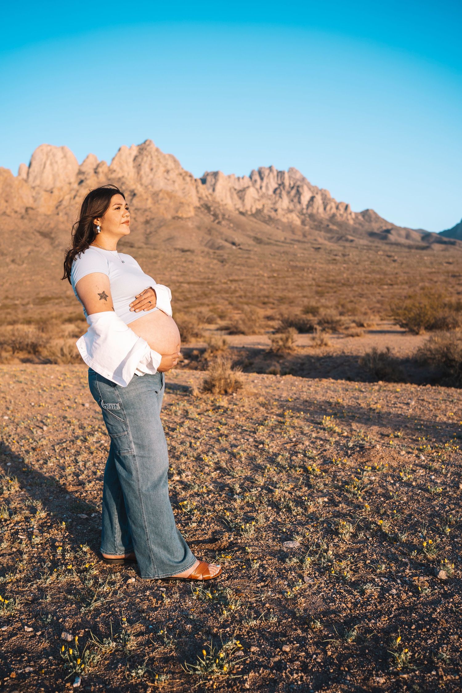 Glowing pregnant woman in white top and jeans stands proudly in desert with rocky mountains behind.