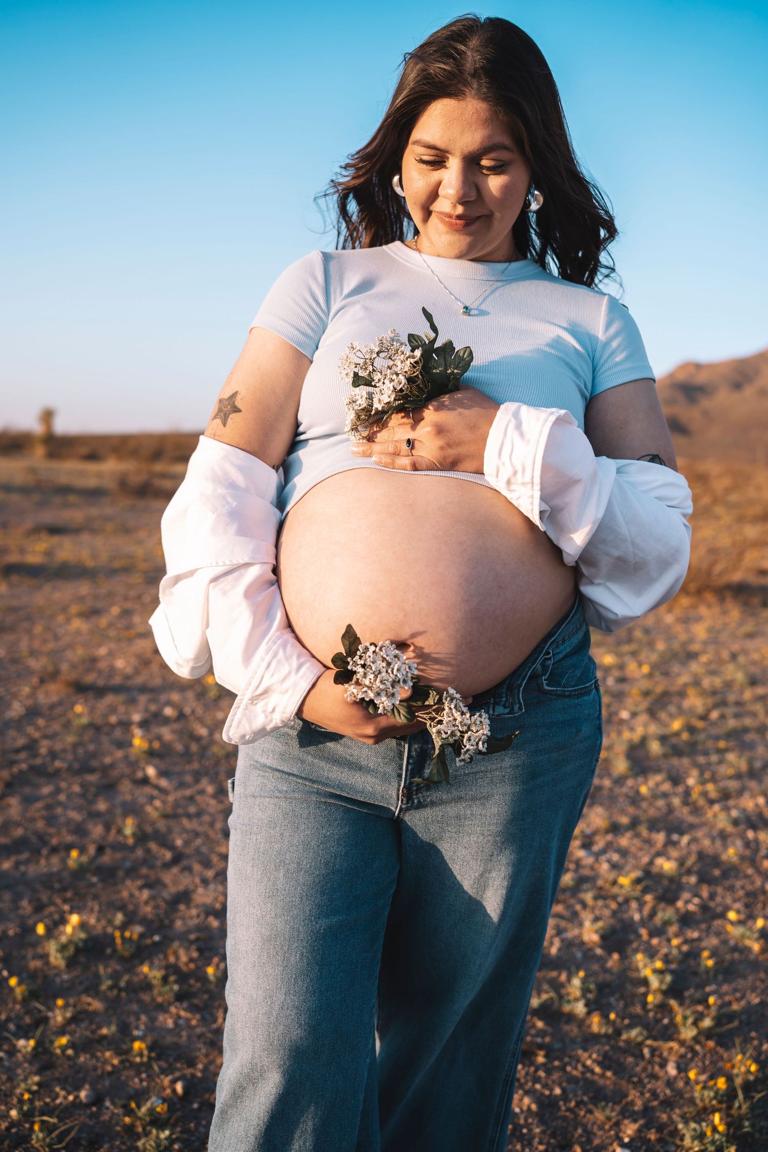 Smiling pregnant woman holding wildflowers against her bare baby bump, wearing a blue top and jeans in a desert setting.