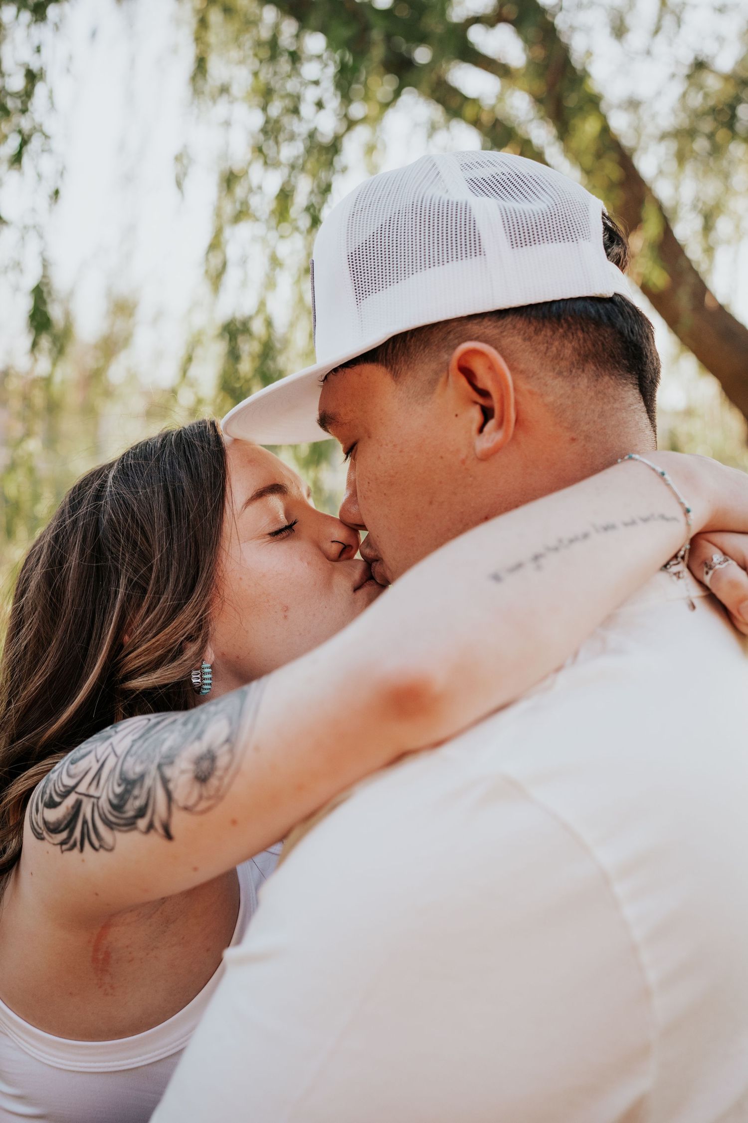 Couple sharing a passionate kiss outdoors, woman with floral arm tattoo embracing man in white trucker hat.