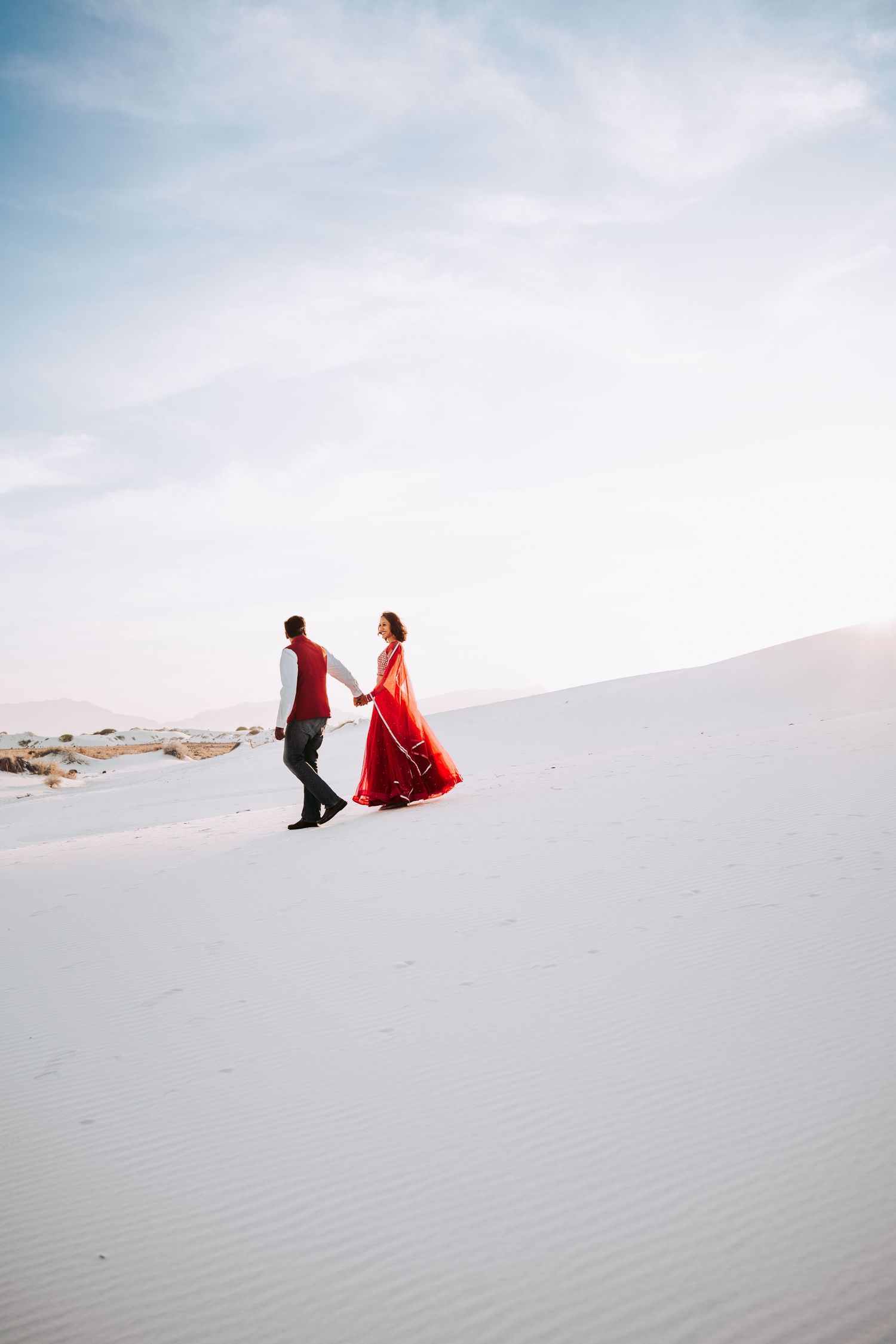 Indian couple in red traditional attire holding hands while walking along white sand dunes.