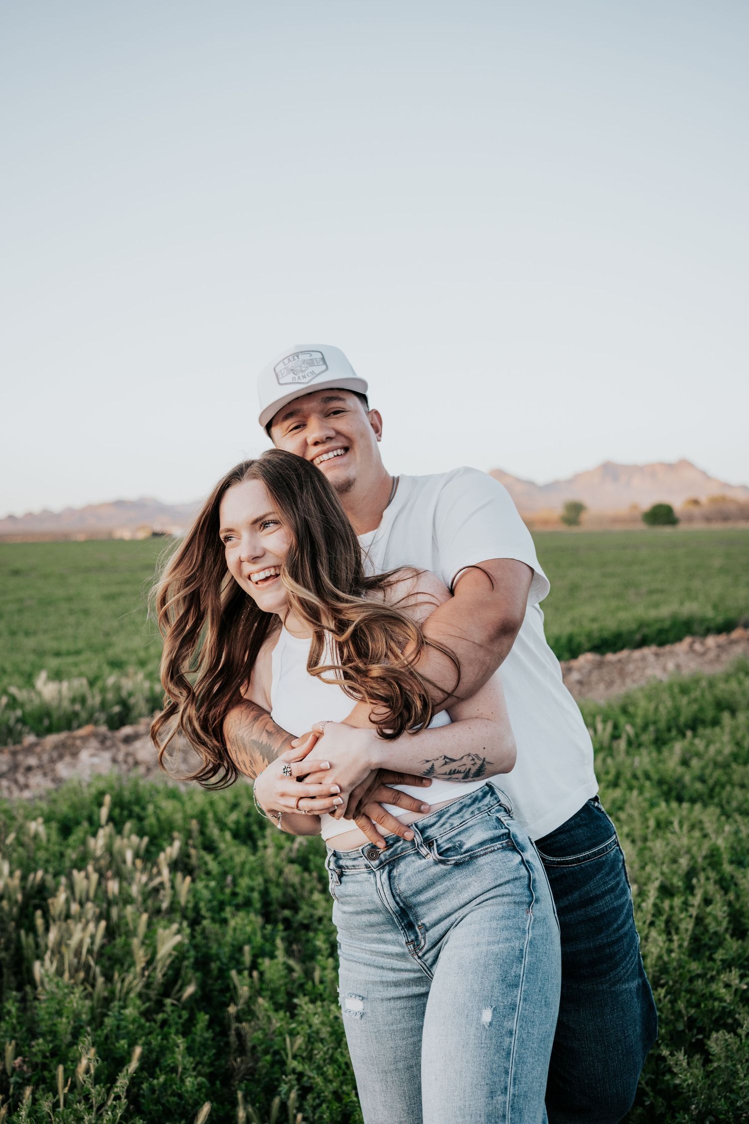 Happy couple embracing in a green field at sunset, man in white cap hugging woman from behind.