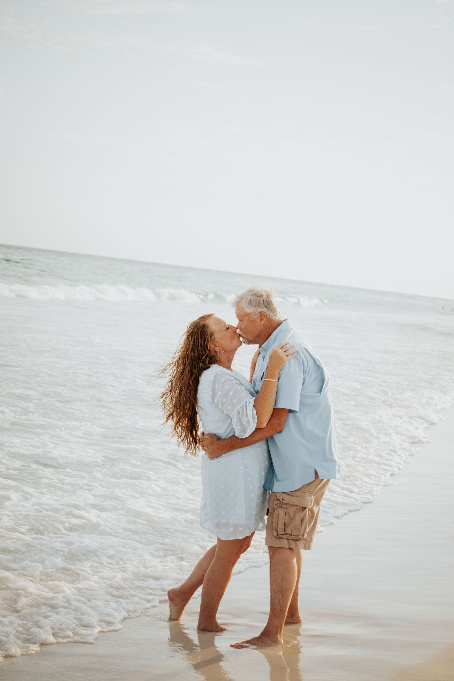 Loving couple sharing a kiss while standing in ocean waves on sandy beach, dressed in coordinating blue outfits.