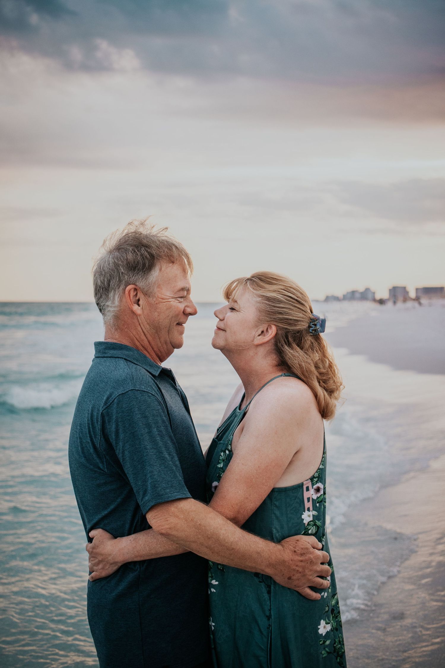 Couple embracing and smiling at each other on a beach at sunset with ocean waves behind them.