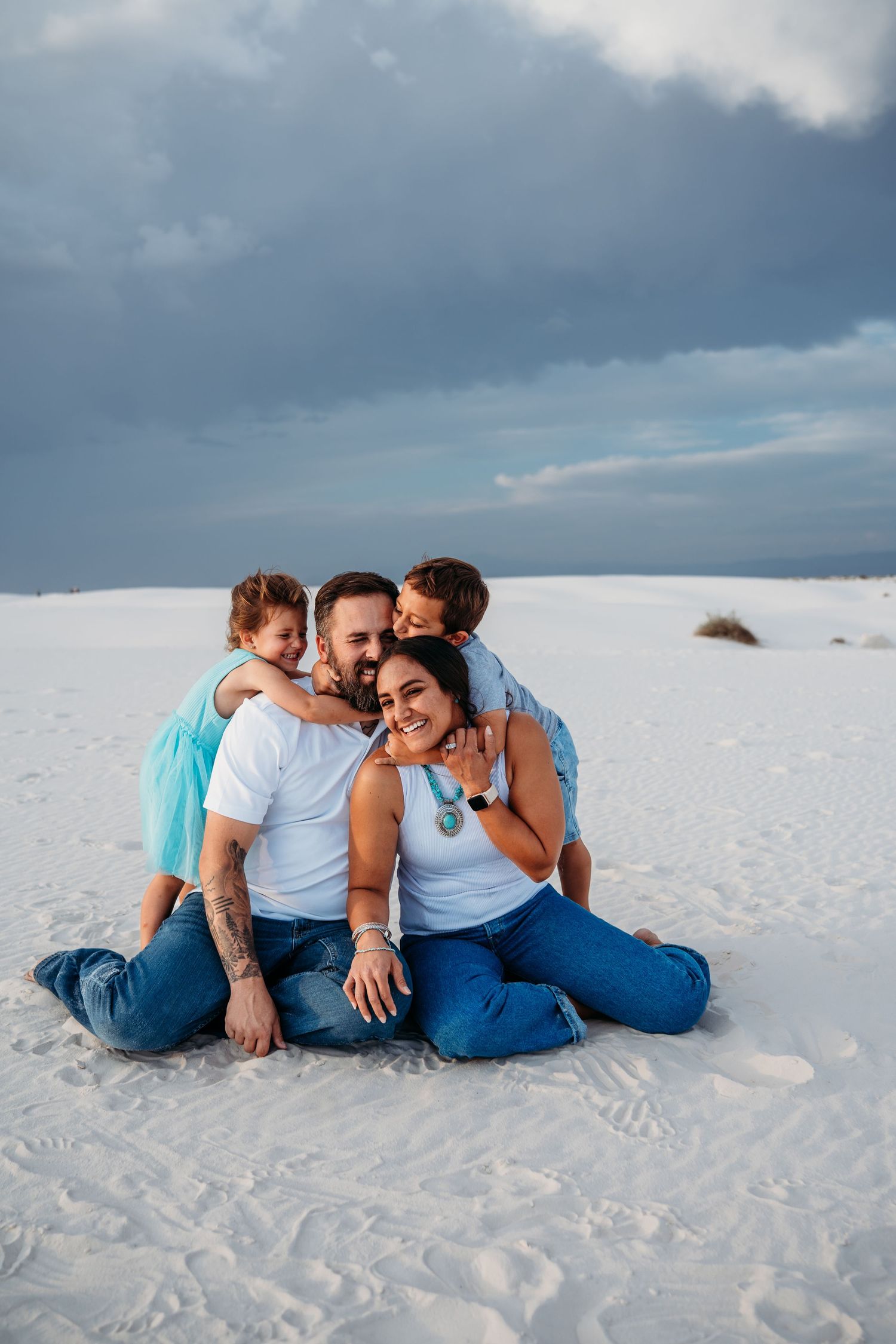 A happy family of four sits together on white sand dunes, with children kissing their parents.