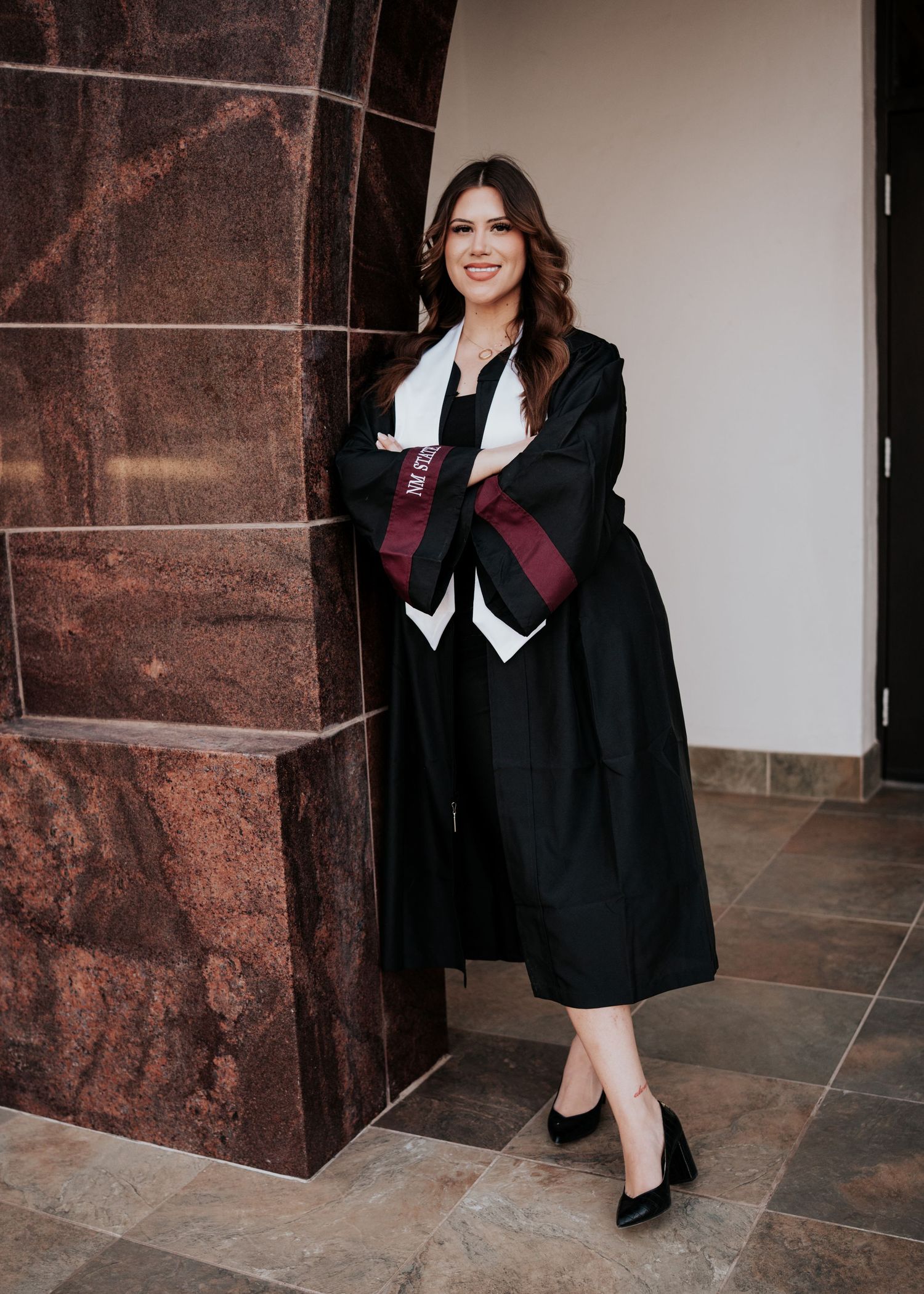 Smiling female graduate in black gown with maroon NM State stole posing by stone pillar.