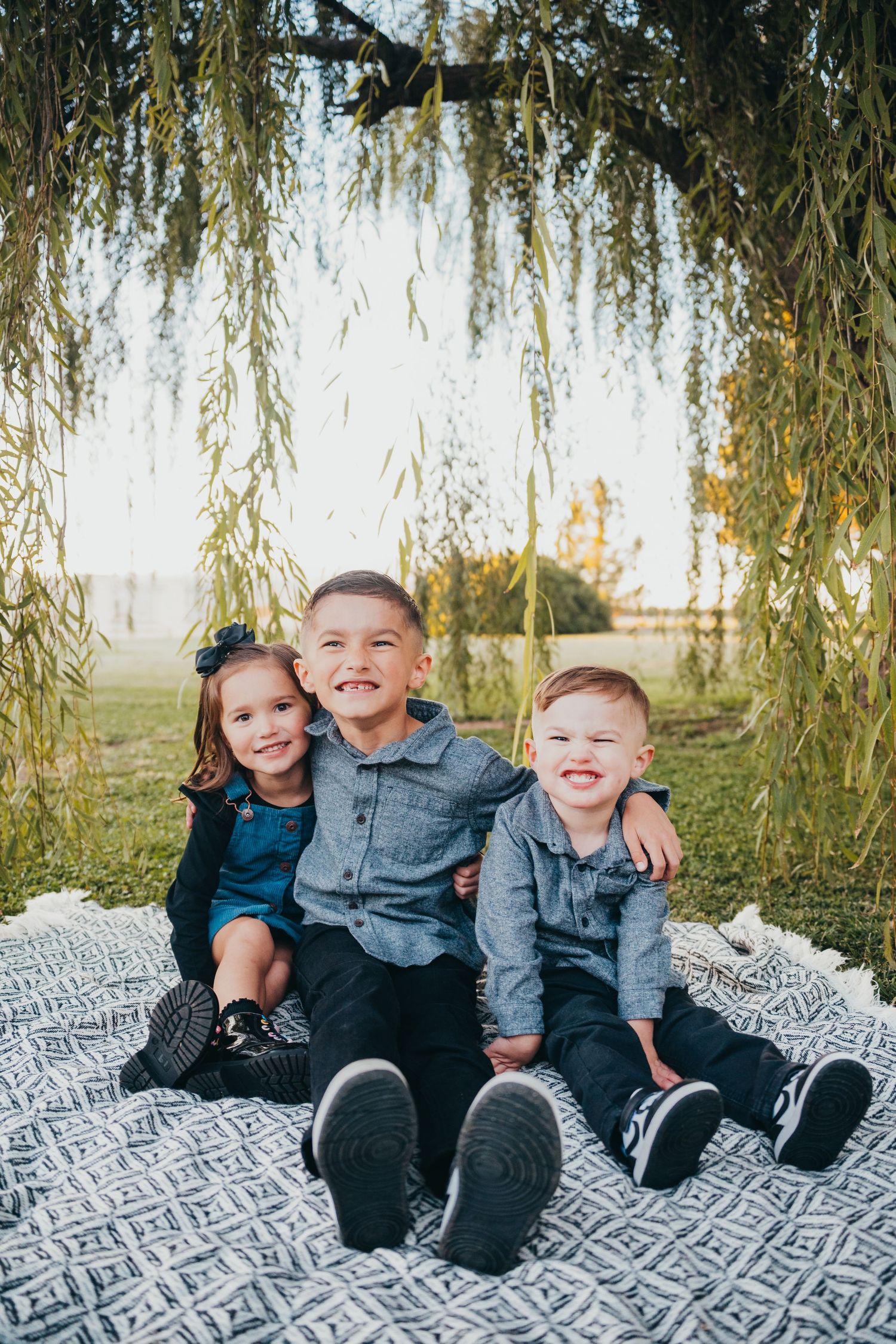 Three young siblings sit together on a blanket under a weeping willow tree, smiling for a family portrait outdoors.