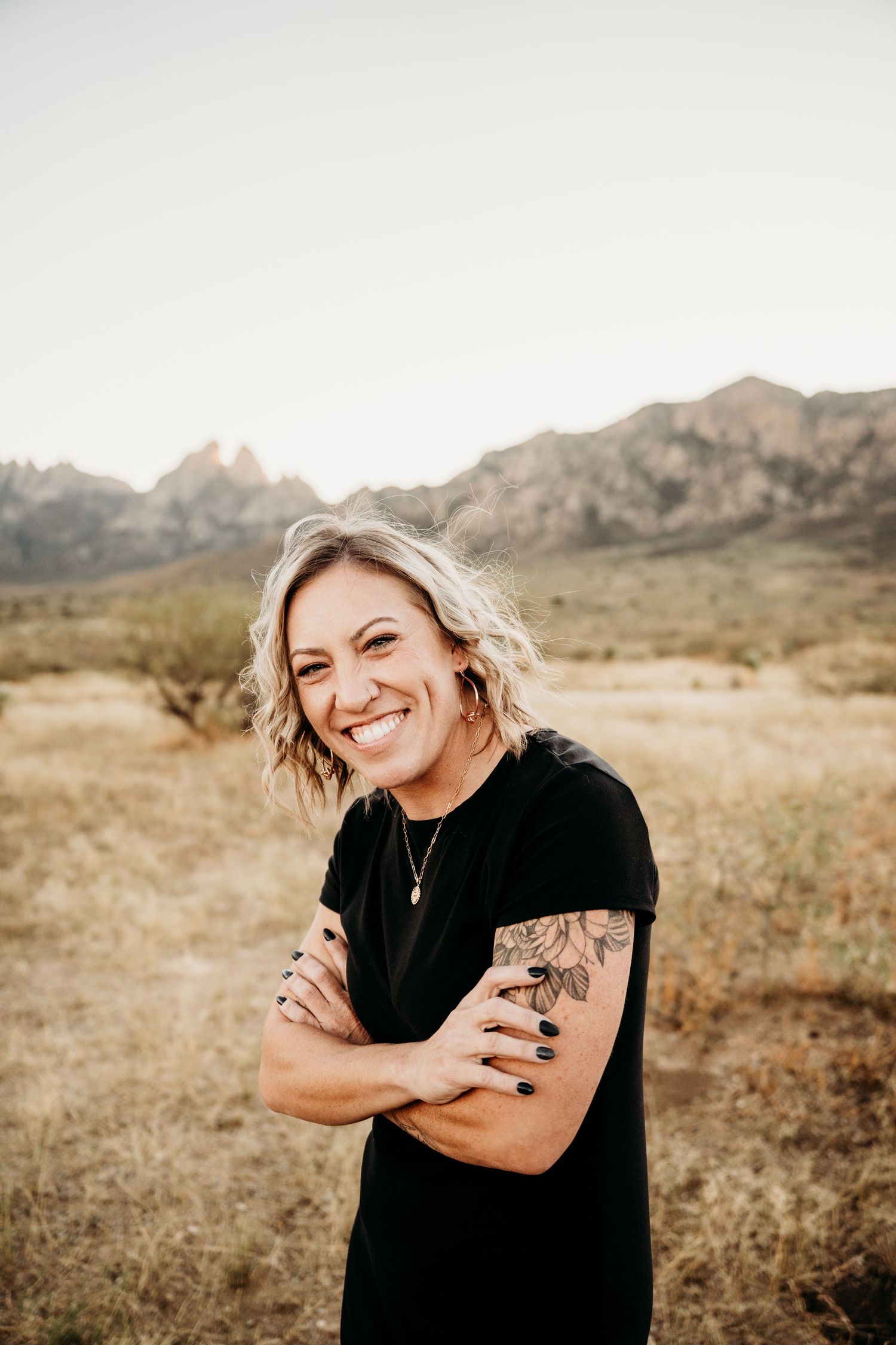 Smiling woman with floral arm tattoo stands arms crossed in golden desert field with mountains.