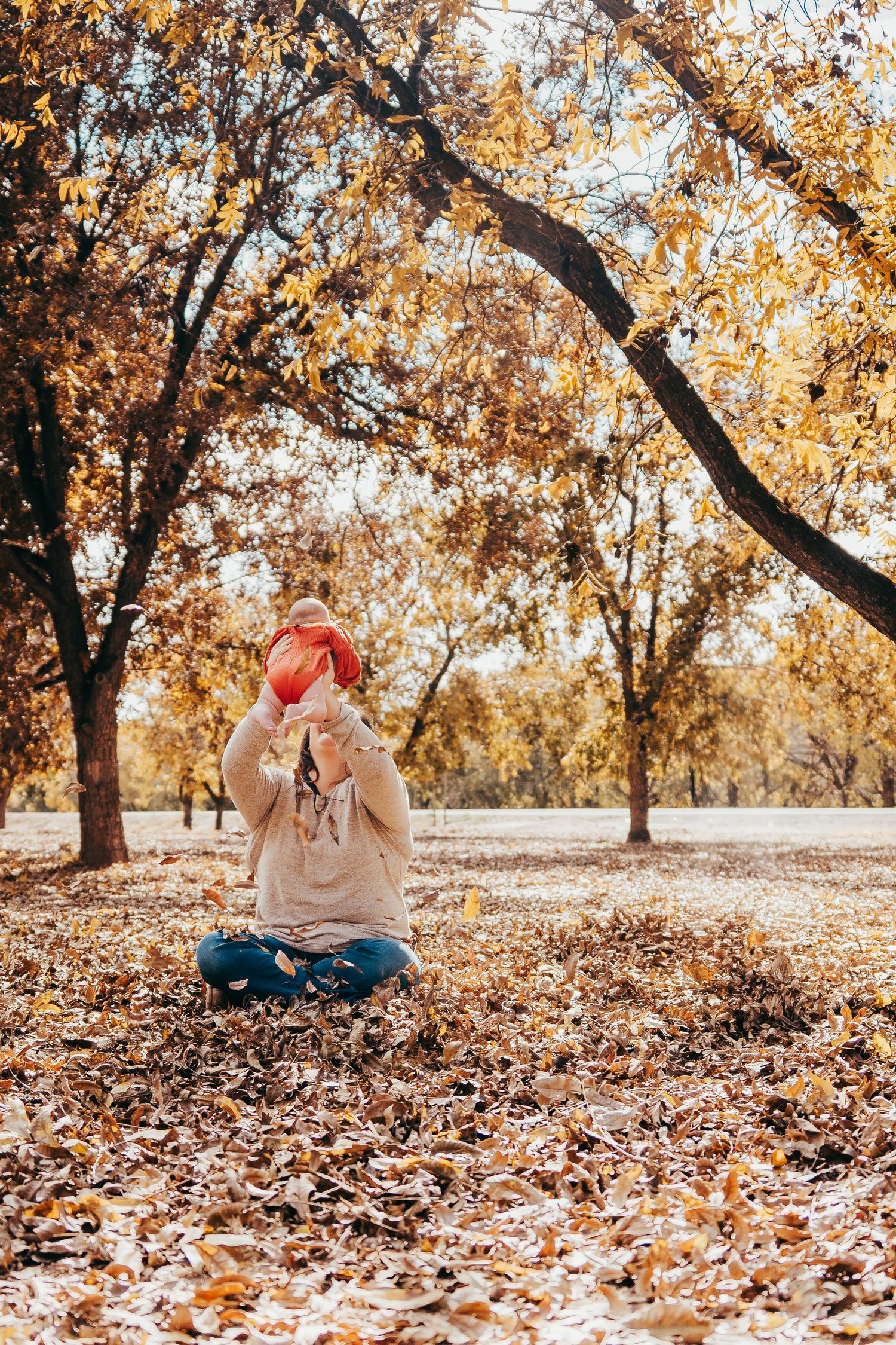 A woman sits cross-legged on fallen autumn leaves holding a baby up to her face outdoors.