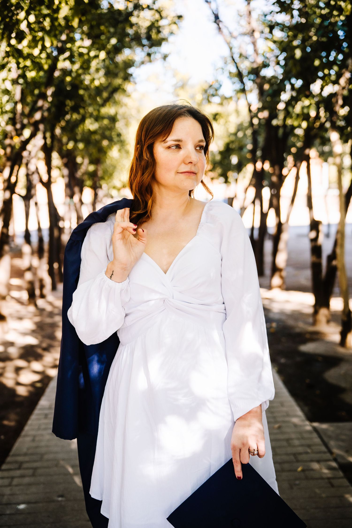 Woman in white long-sleeve dress holding navy blazer poses on tree-lined pathway in warm sunlight.