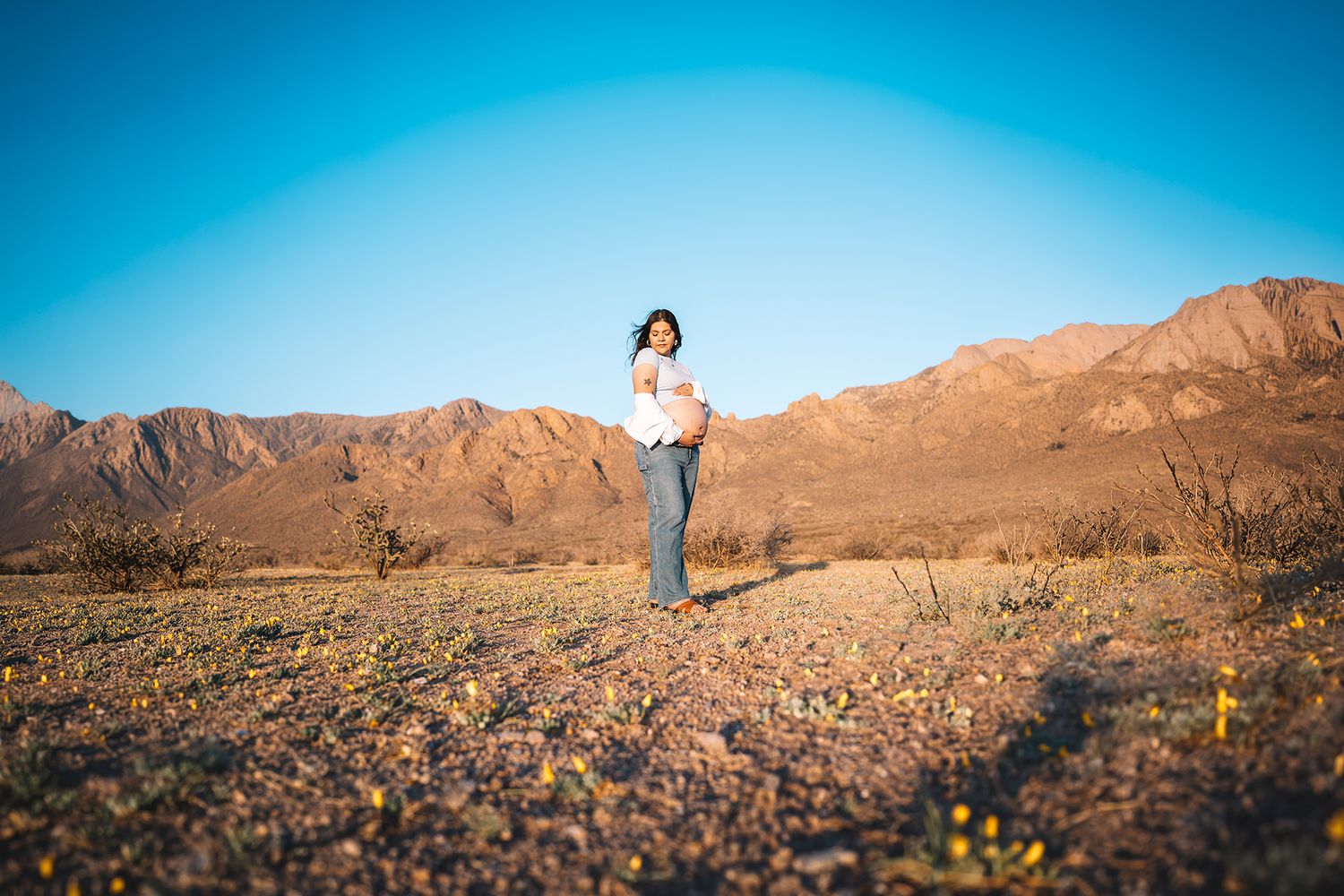Pregnant woman stands in desert field with mountains and blue sky behind her.