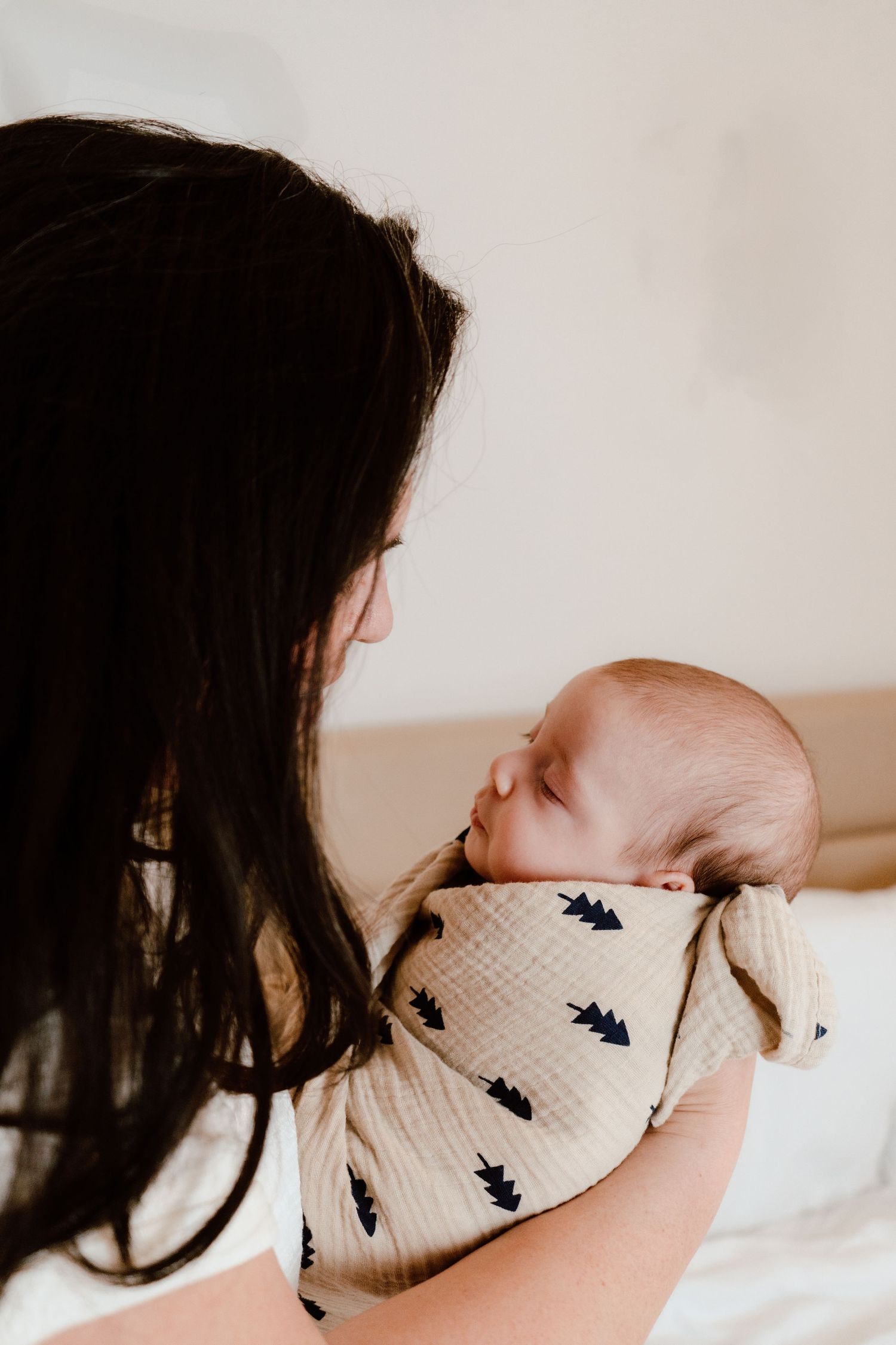 Dark-haired mother holding sleeping newborn baby wrapped in cream muslin swaddle blanket indoors.