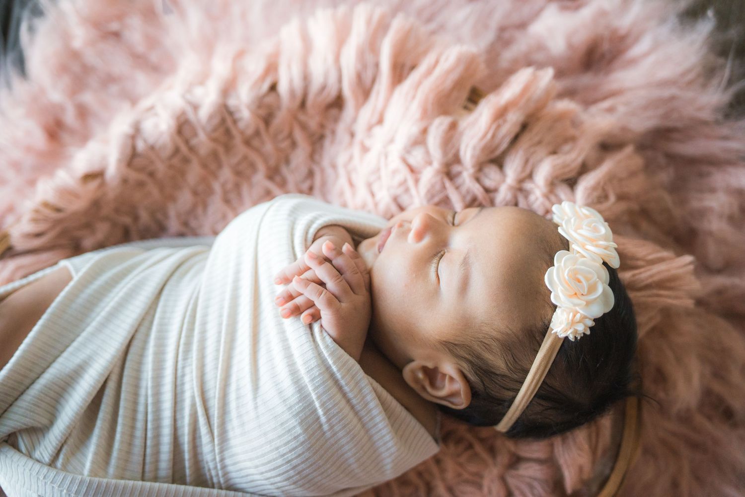 Newborn baby girl sleeping peacefully swaddled in white wrap with cream floral headband on pink fur.