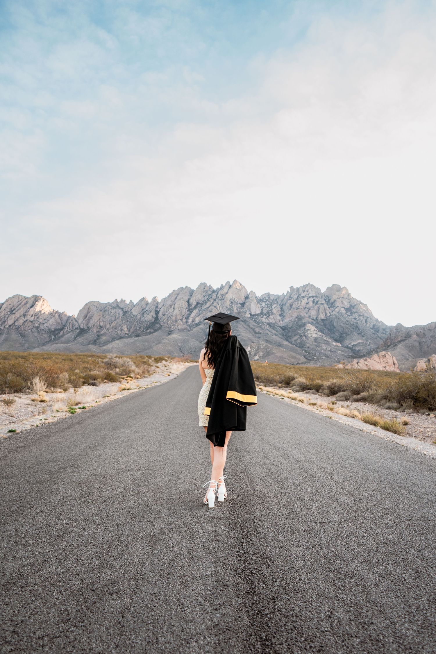 Female graduate in black and gold cap and gown walks away down a scenic desert road toward rocky mountains.