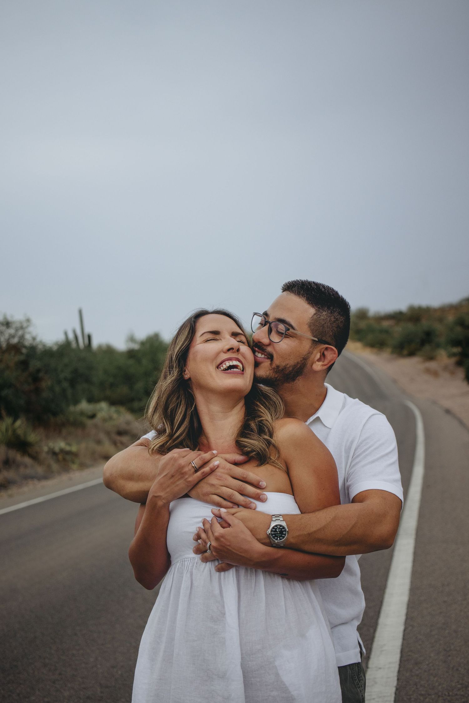 Couple embracing on desert road, man kissing woman's cheek as she laughs joyfully in white dress.