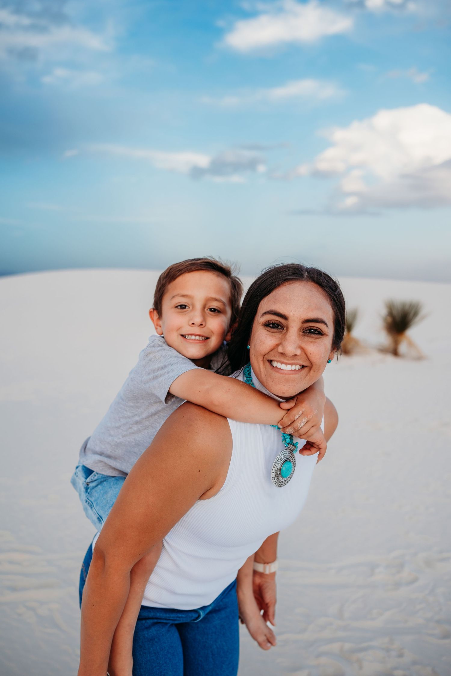 Smiling mother giving young boy a piggyback ride on white sand dunes under blue sky.