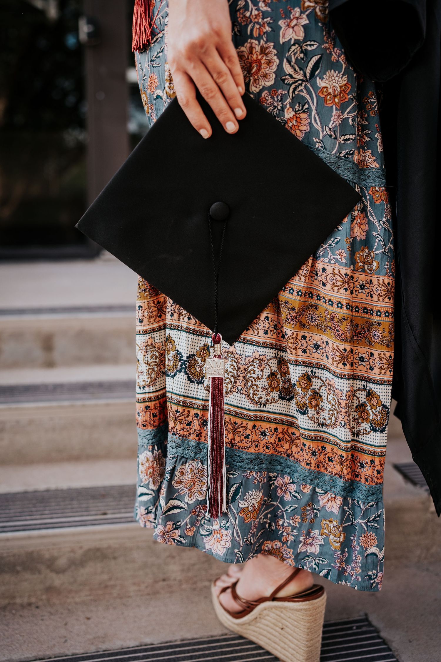 Close-up of graduate holding black mortarboard cap in floral dress with wedge sandals.