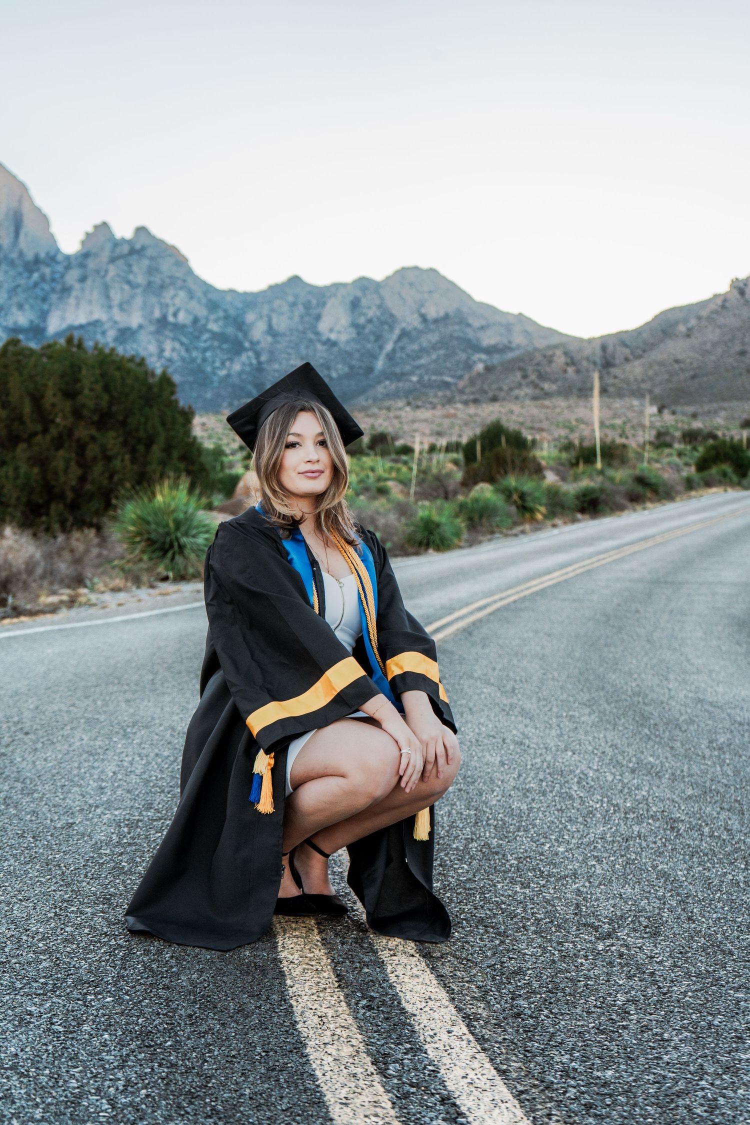 Graduate in black cap and gown crouching on road centerline with desert mountains in background.