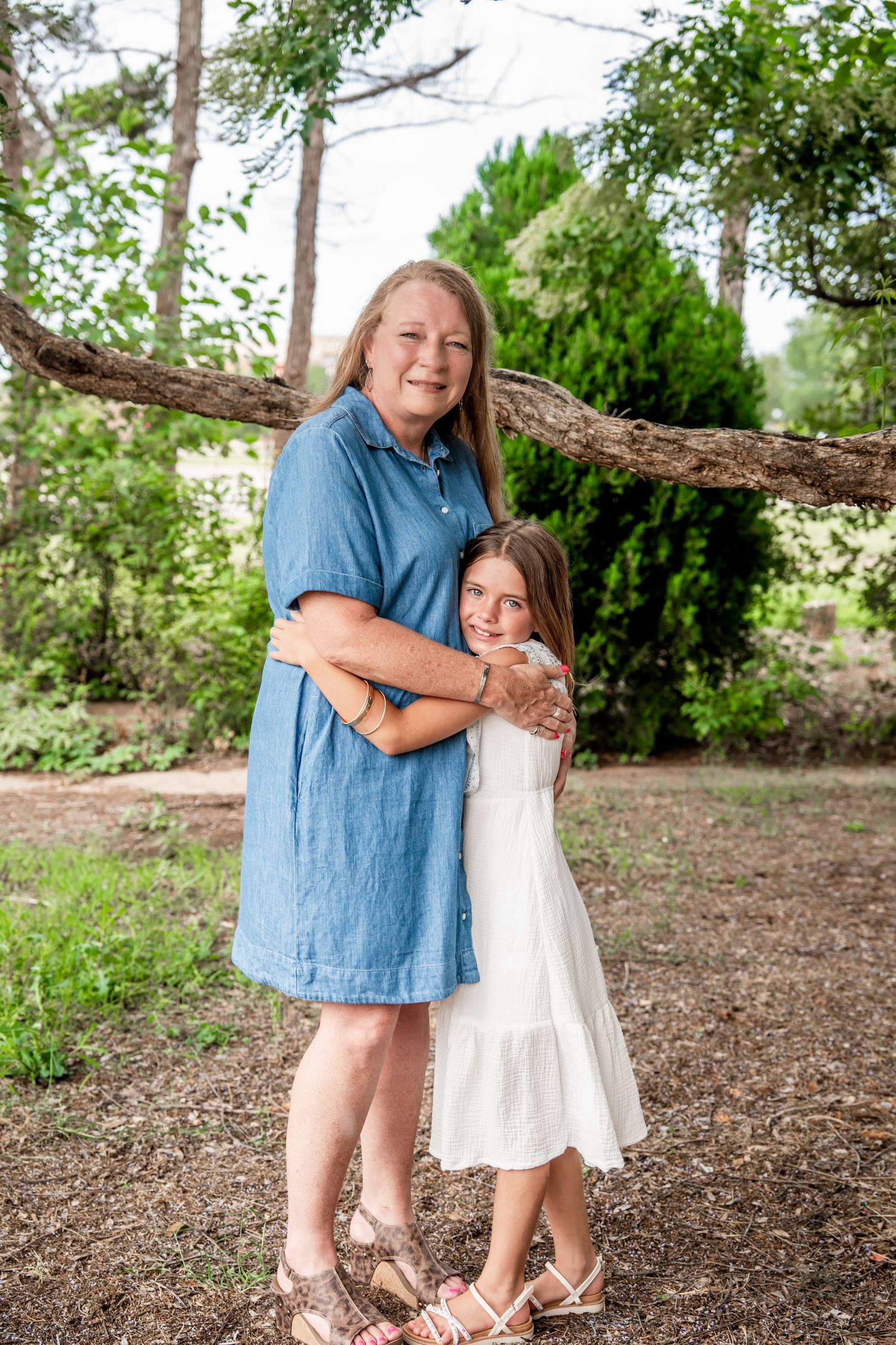 Older woman in blue dress embracing smiling girl in white dress outdoors near trees.