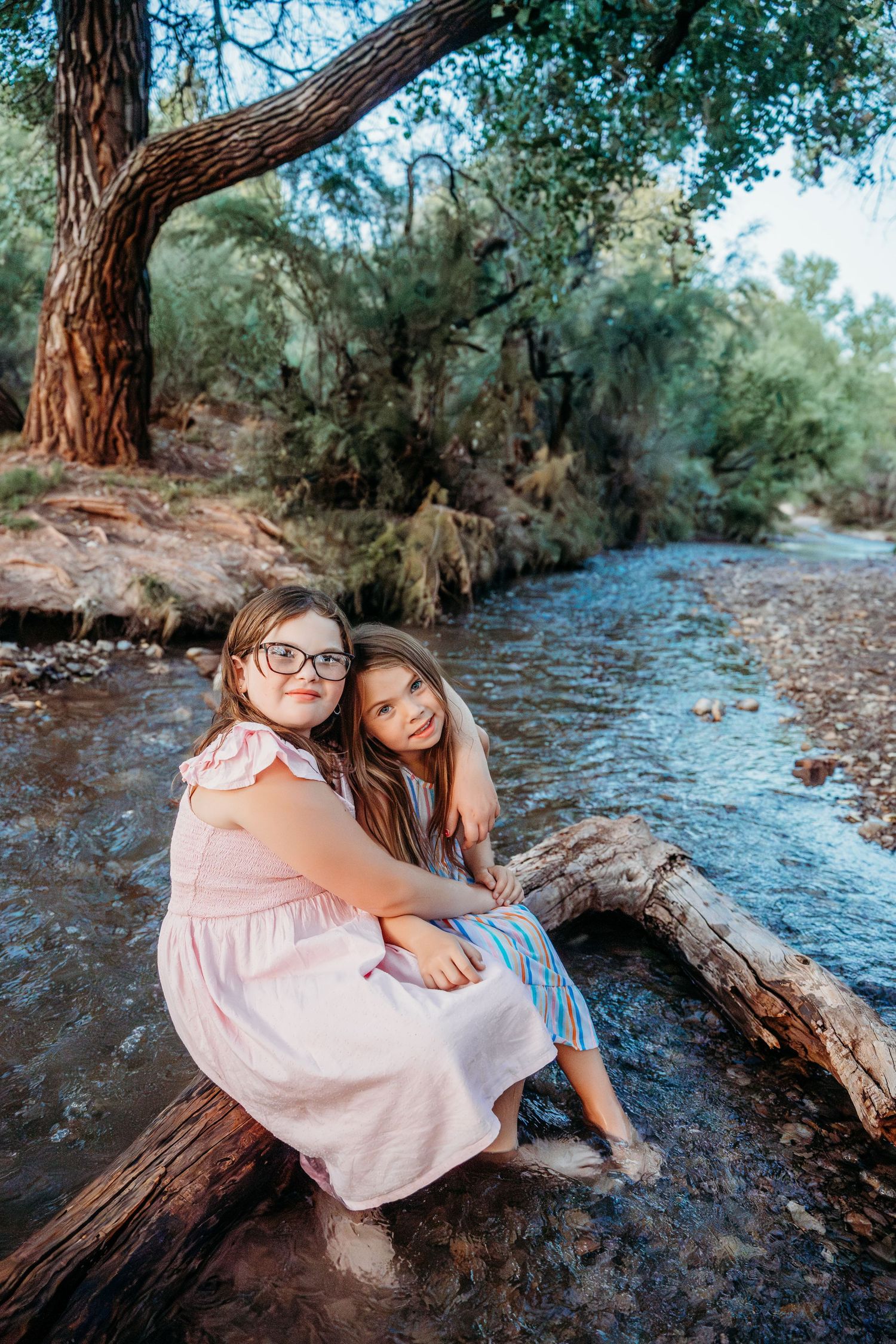 Two girls sit together on a log by a peaceful creek, embracing and smiling warmly.