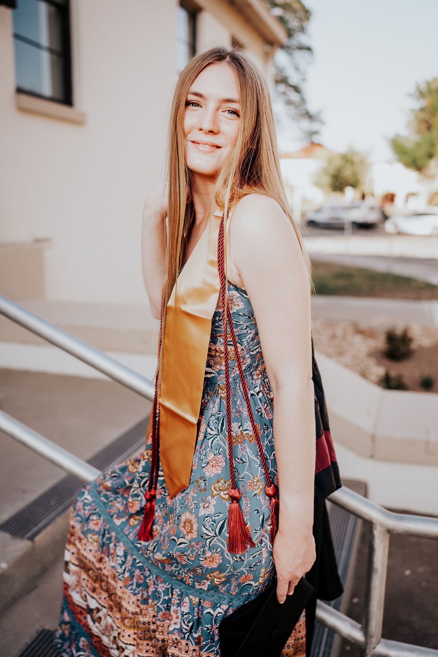 A smiling female graduate in a floral dress wearing a gold and maroon graduation stole poses on sunny steps.