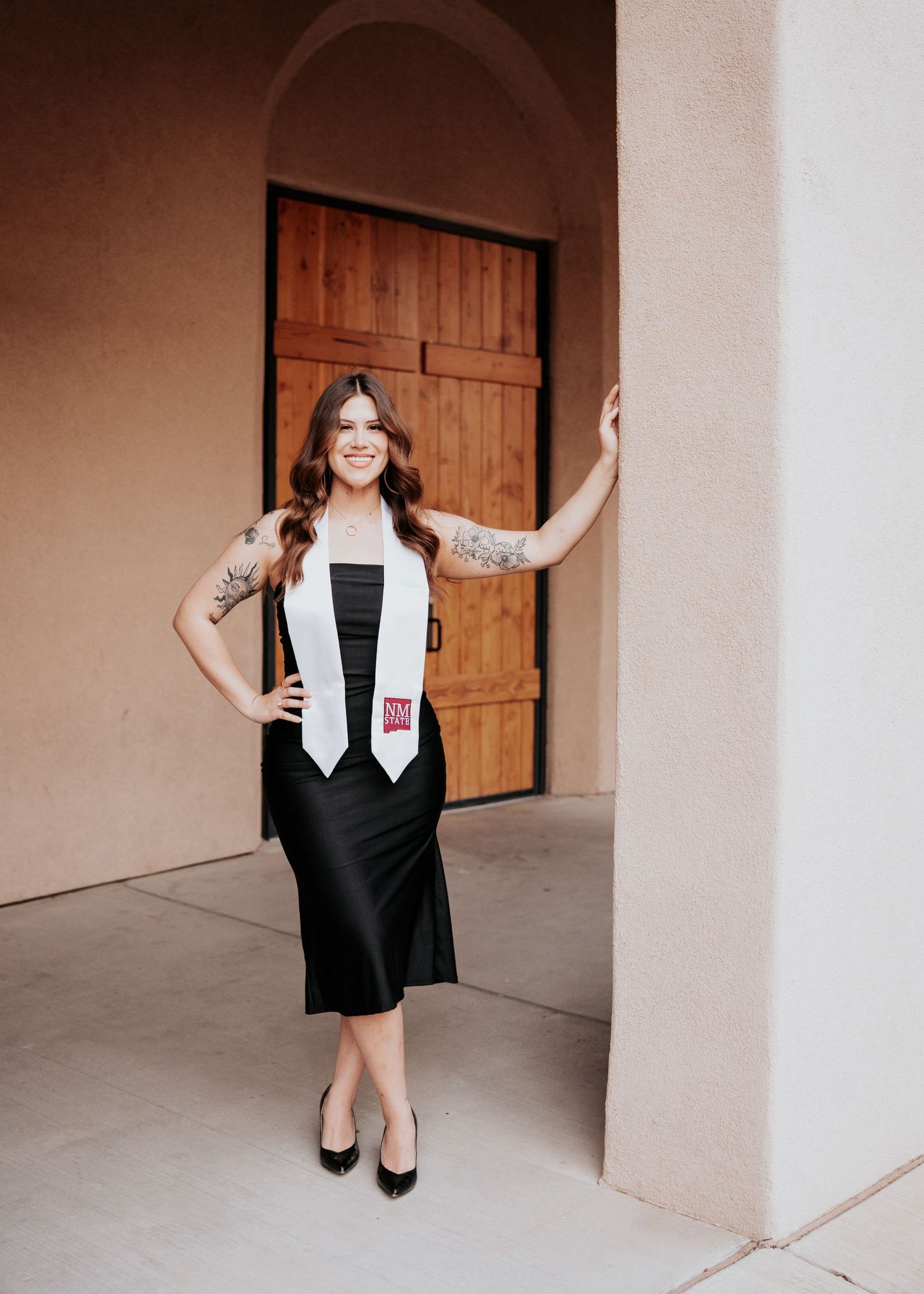 Smiling graduate in black dress with white NM State stole posing at adobe building entrance.