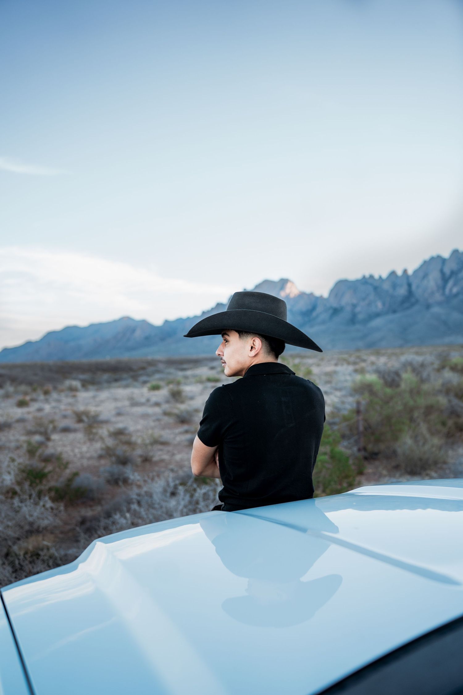 Man in black cowboy hat leaning on white truck gazing at desert mountain landscape.