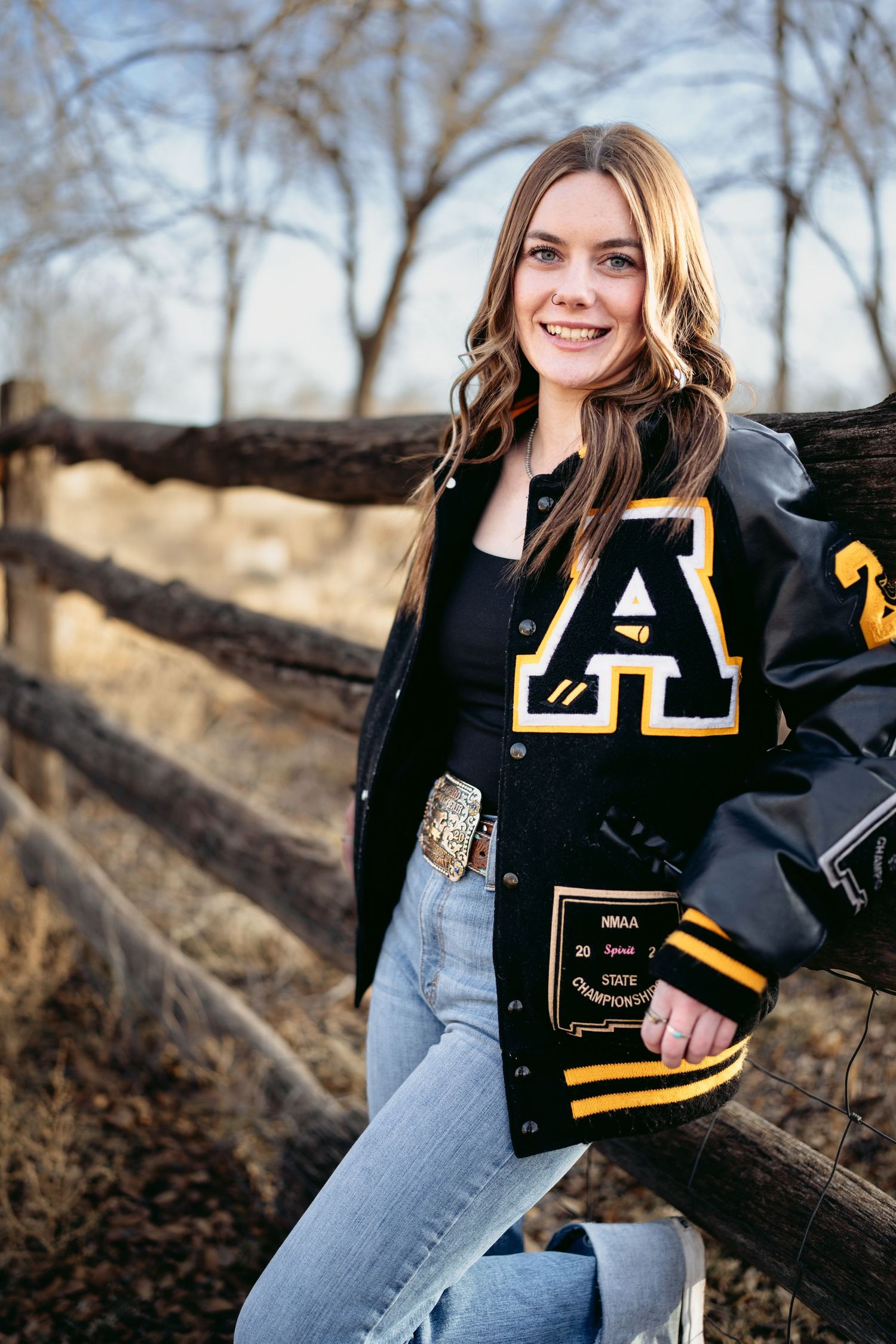 Smiling high school senior girl wearing black and gold letterman jacket leaning against rustic fence outdoors.